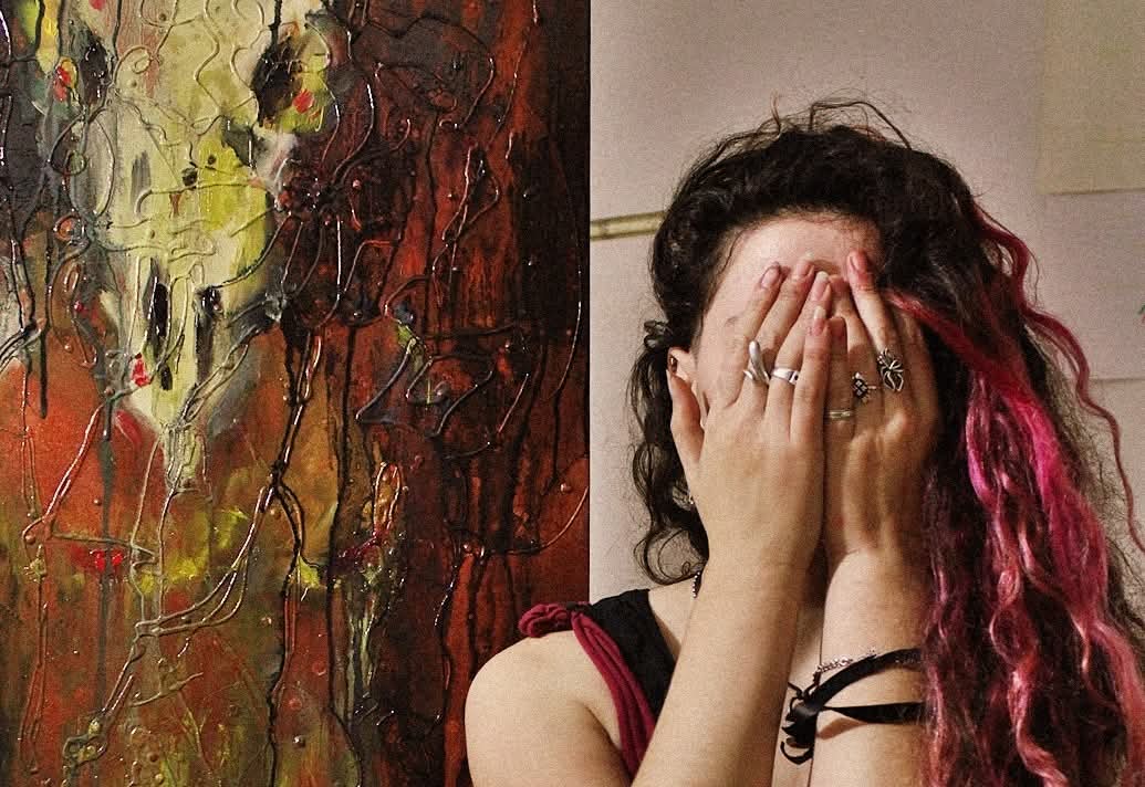 Person with curly hair covering face with hands, pink streaks in hair, wearing rings and bracelets, standing next to abstract painting.