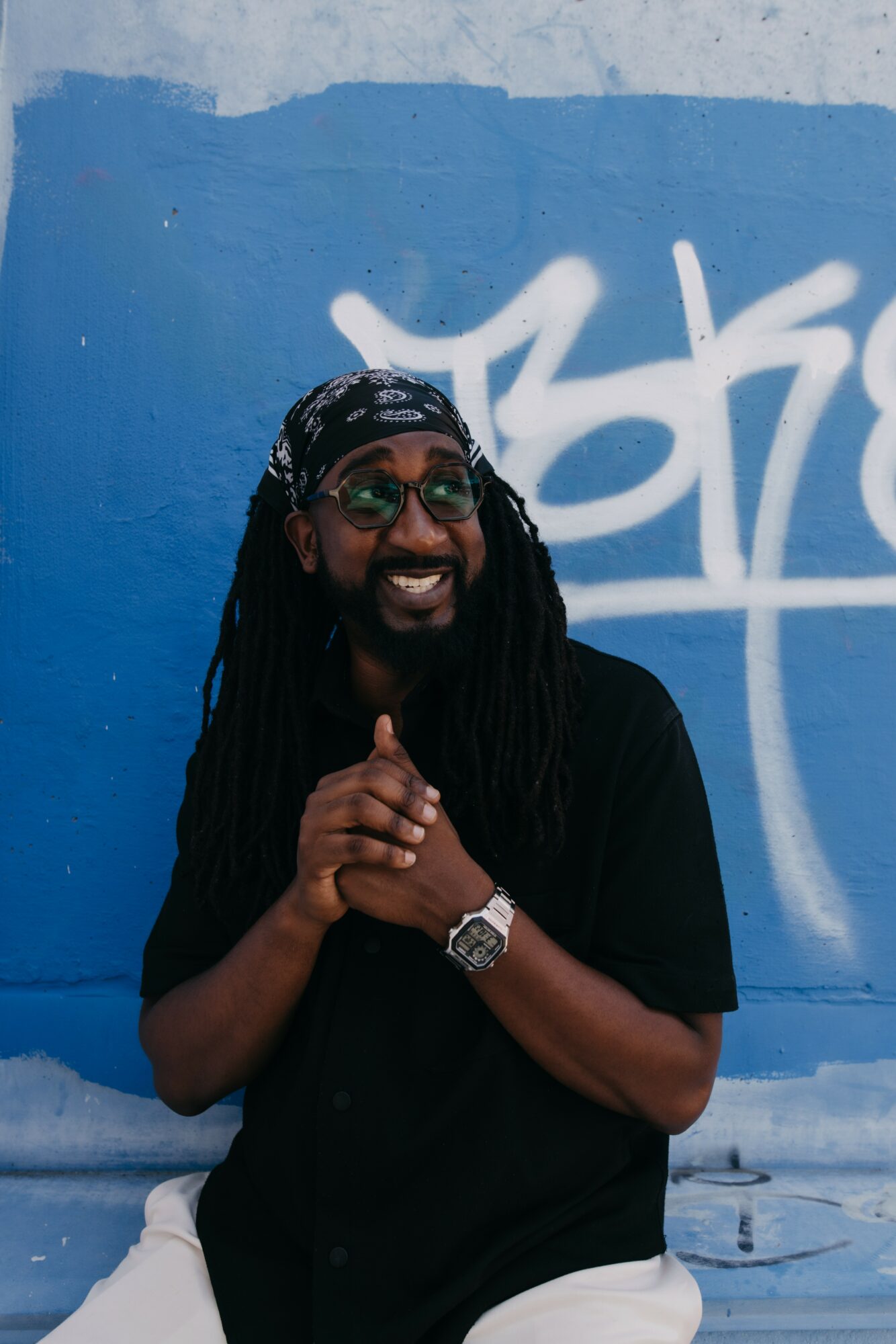 Person with long dreadlocks, sunglasses, and a bandana, smiling and clapping hands in front of a blue wall with graffiti.