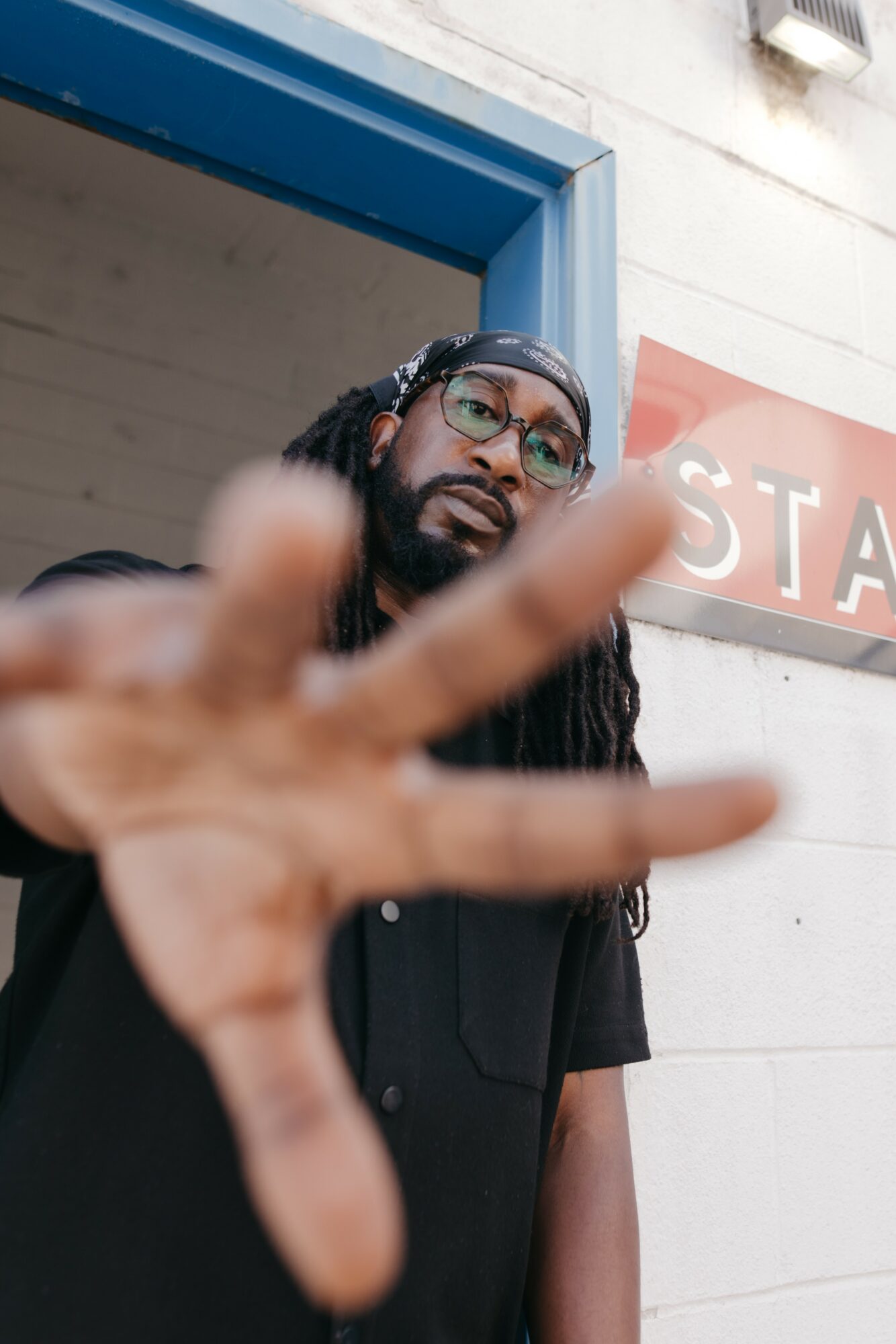 Man with glasses and dreadlocks making a peace sign close-up, background of blue door and white wall.