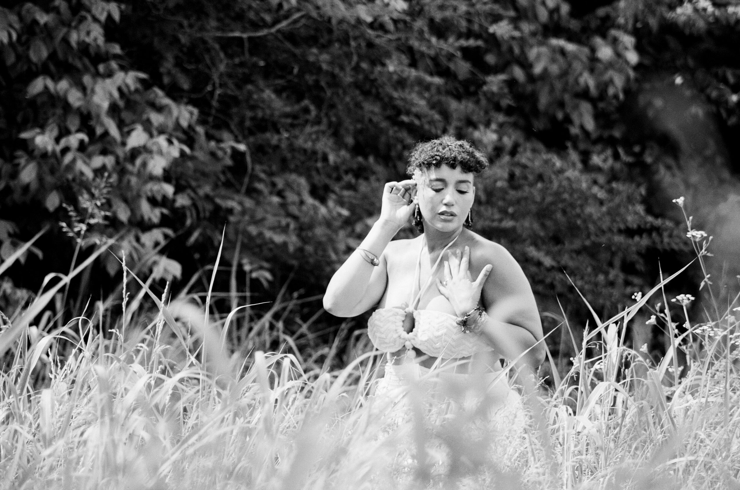 Woman with curly hair in a field of tall grass, touching her face and chest, with trees in background.