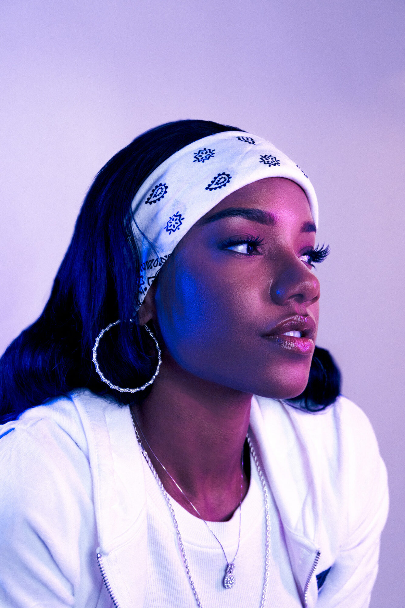 Young woman with dark hair wearing a white headband and hoop earrings, looking to the side against a purple background.
