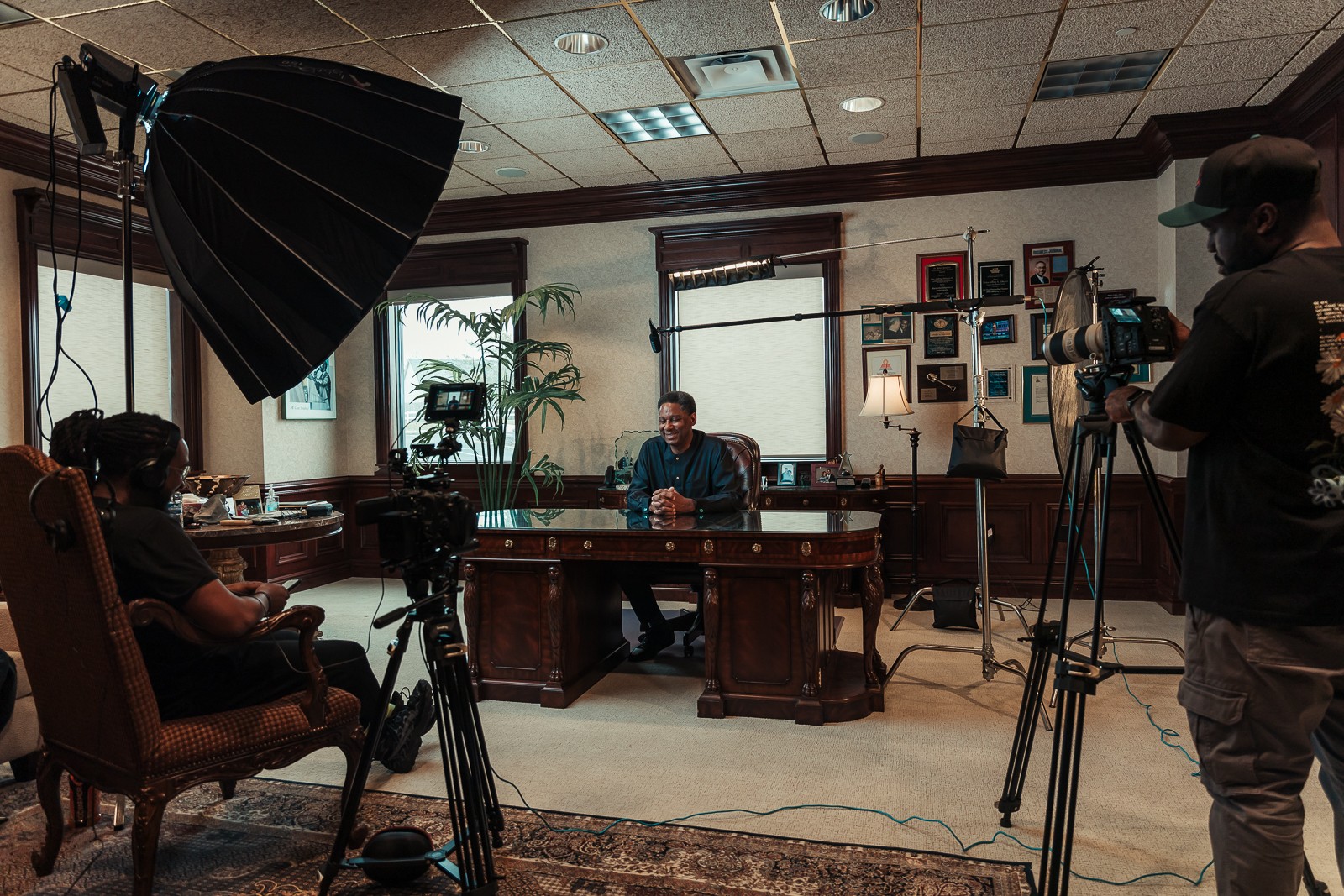 Person sitting at a desk in a room with cameras and crew filming a man at the desk, with windows and plants in background.