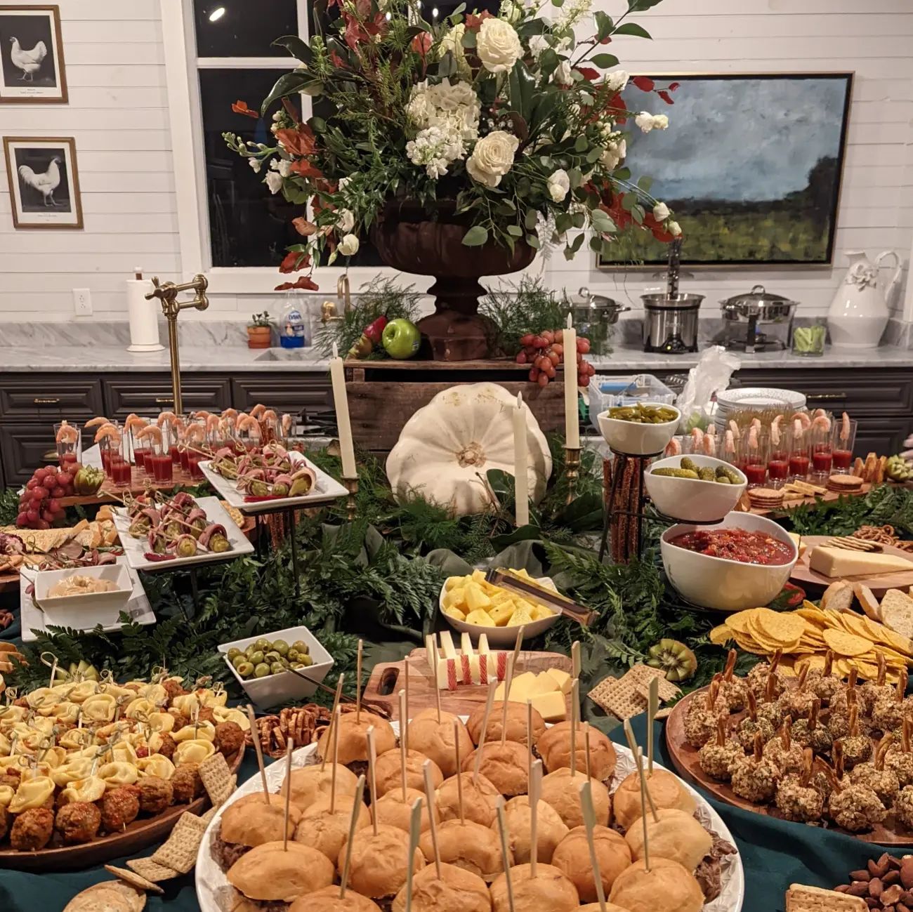 Table with various appetizers, bowls, and a large floral centerpiece in a decorated room.