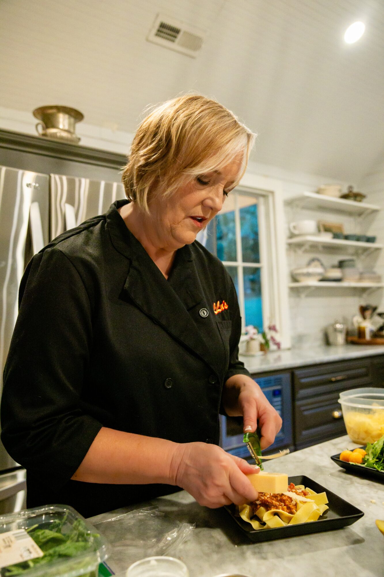 Woman with short blonde hair preparing food in a kitchen, wearing a black chef's coat, standing at a counter with ingredients.