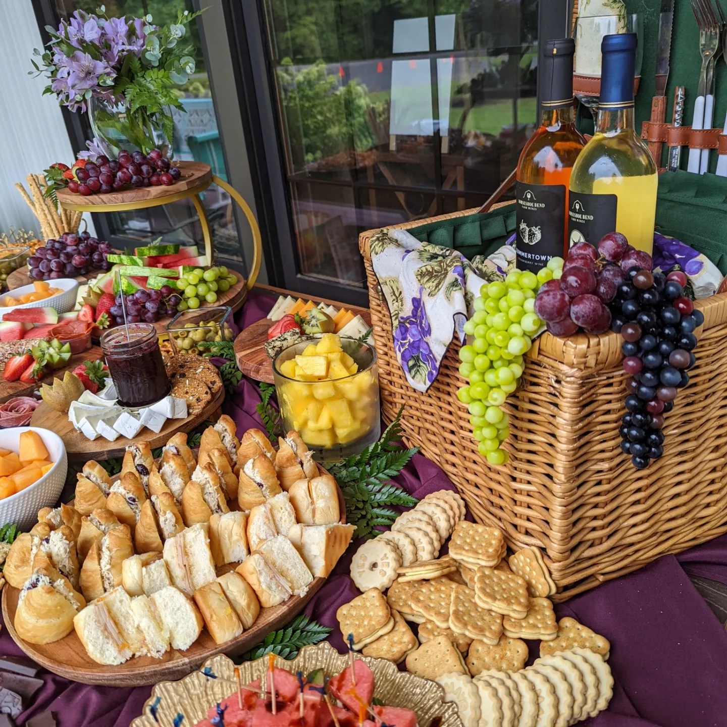 Table with bread, fruit, wine bottles, and grapes near a window, outdoor view visible, purple tablecloth, basket, and flowers.