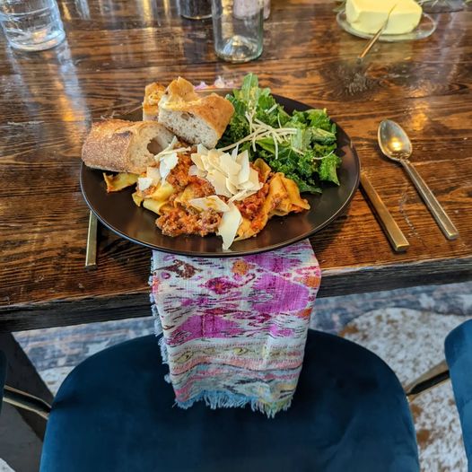 Black plate with salad, bread, and toppings on a wooden table, with a spoon and a colorful cloth hanging from the chair.