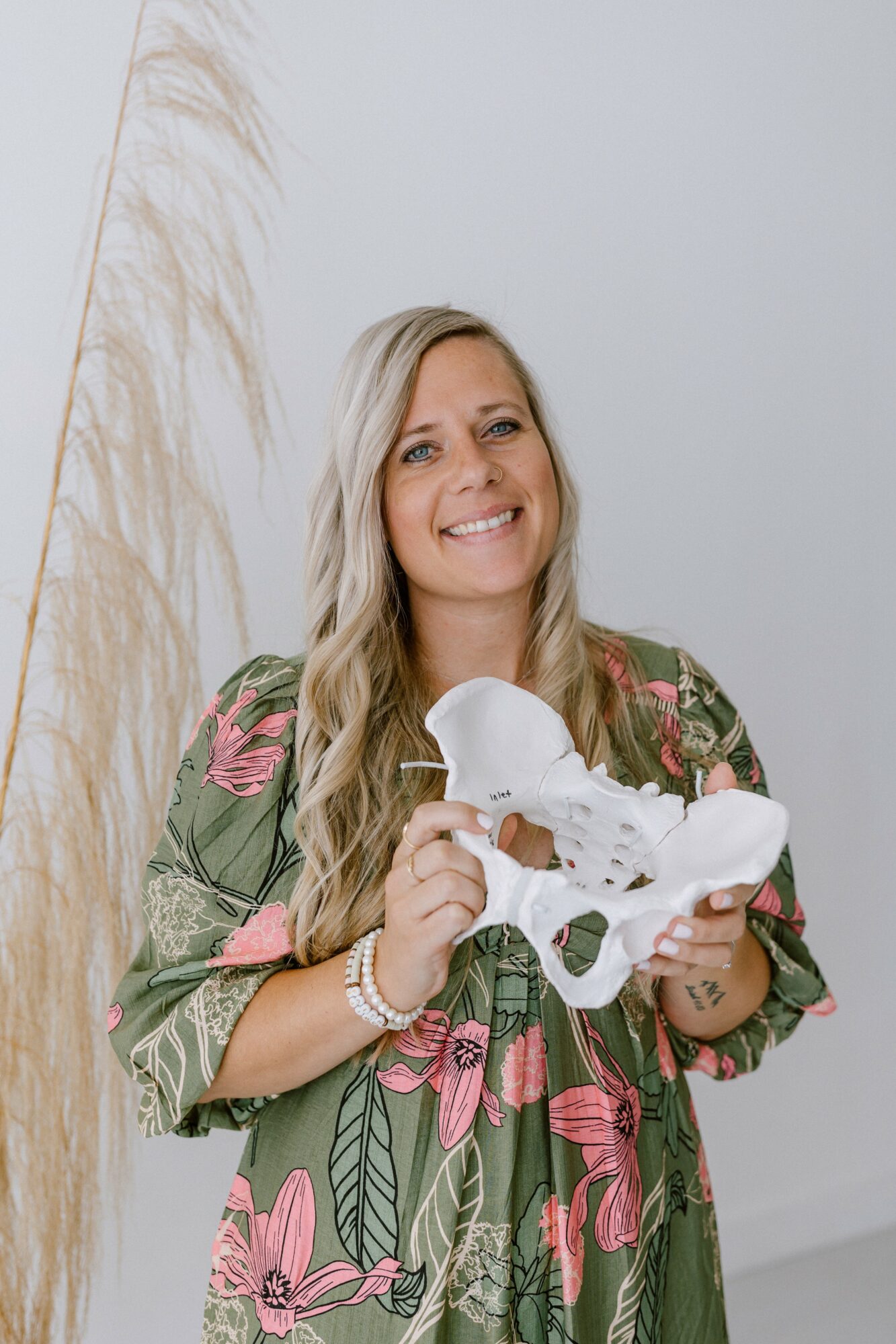 Woman with long blonde hair smiling, holding a white skull model, wearing a floral dress with pink and green patterns.