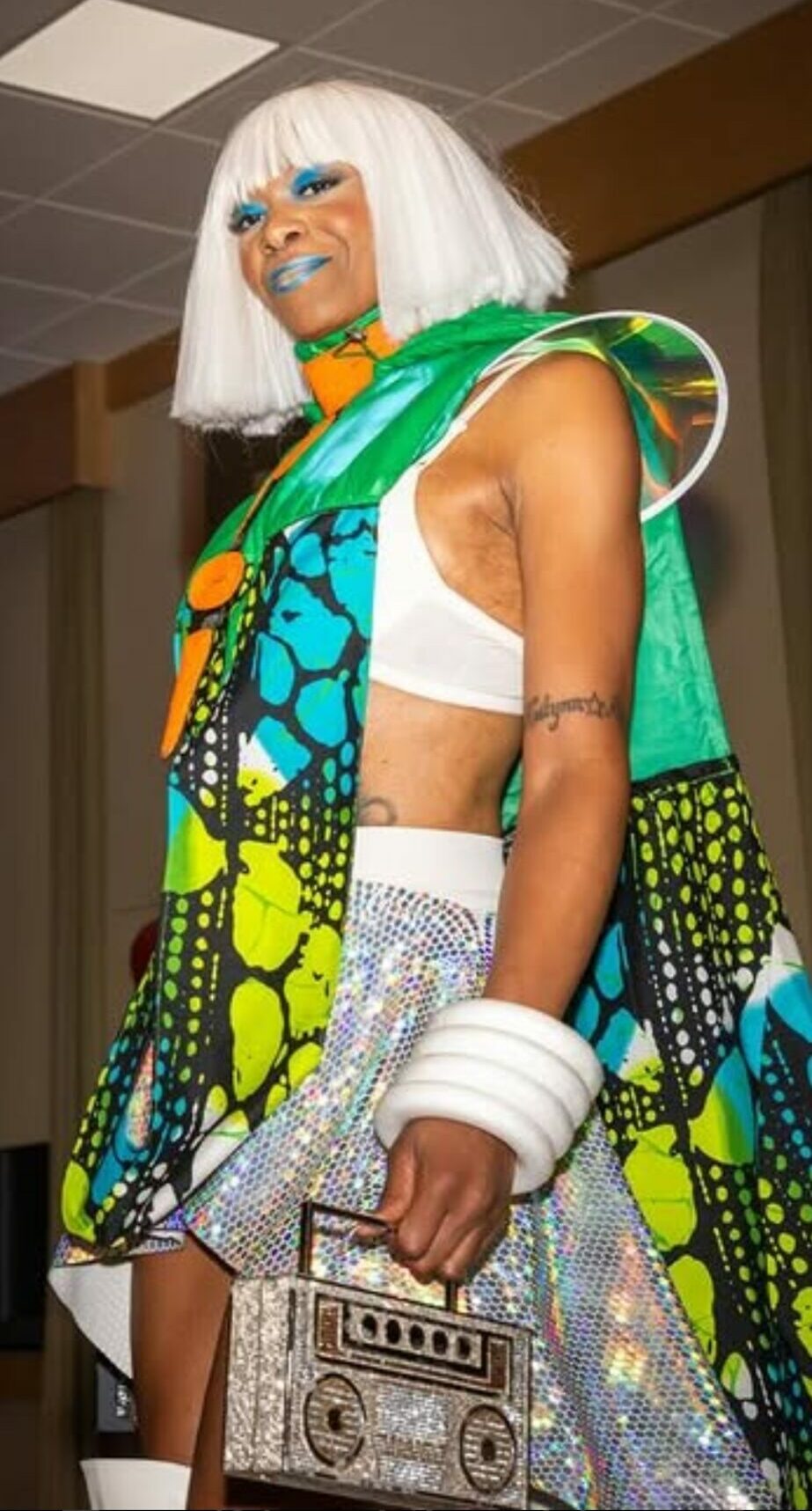 Person with white hair wearing colorful, patterned clothing and sunglasses, holding a silver handbag, smiling.
