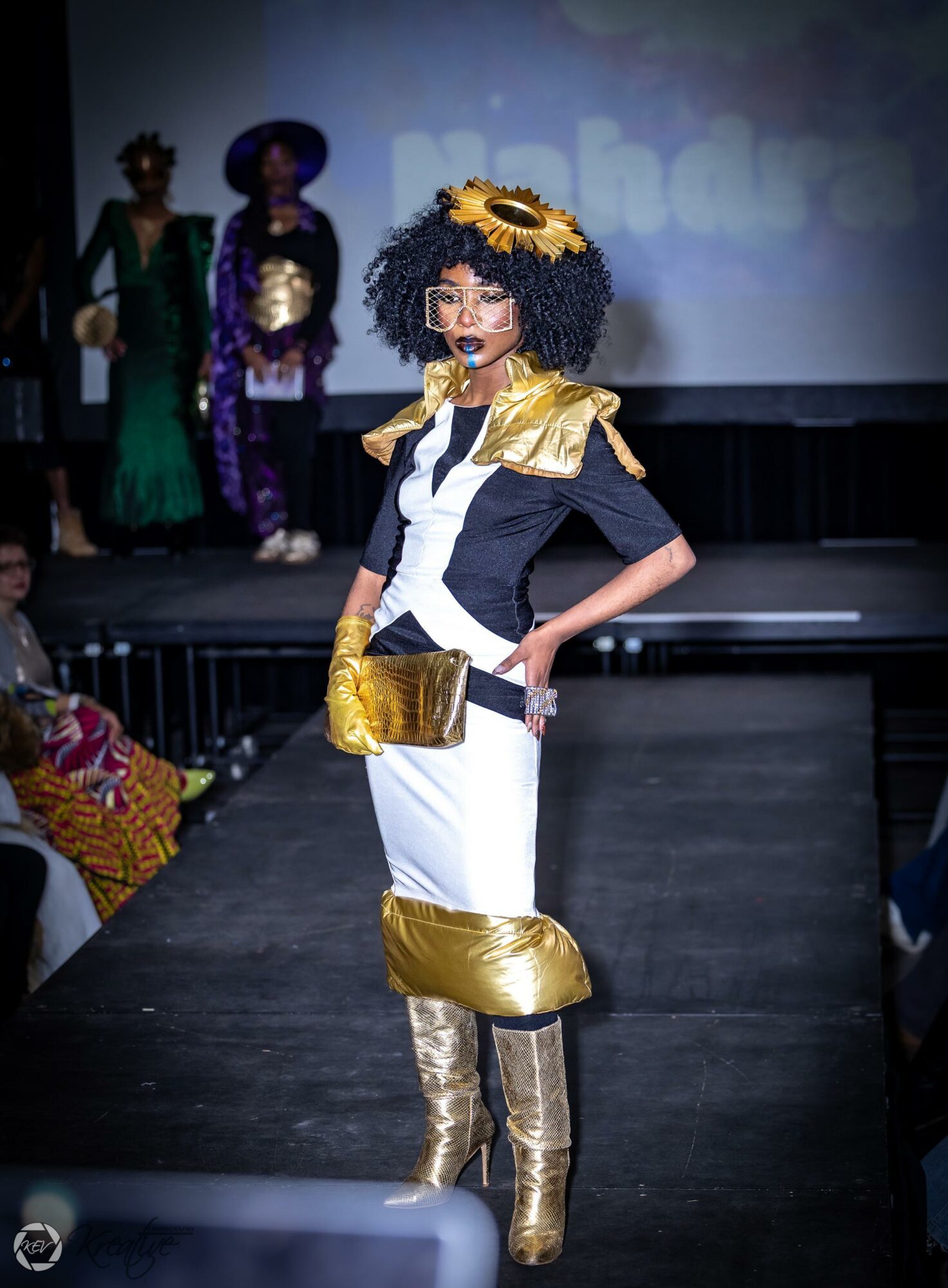 Model in gold and white outfit with gold accessories and boots walks on runway, with two people in costumes in background.