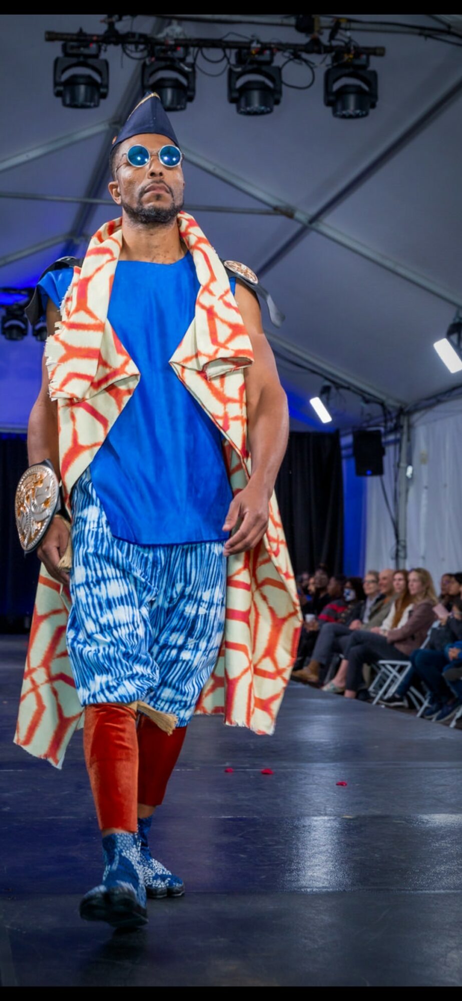 Male model walking on runway wearing sunglasses, a blue dress, patterned coat, and red boots, with audience in background.