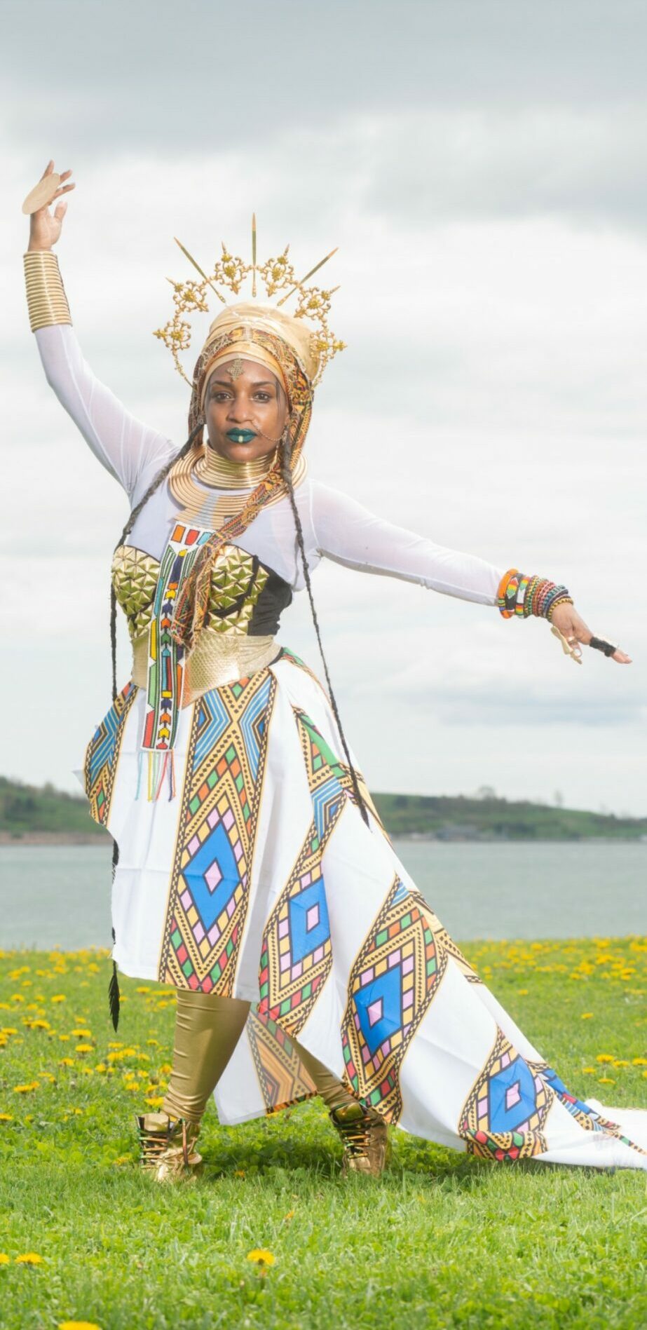 Person in colorful traditional costume standing outdoors with arms raised, cloudy sky background.