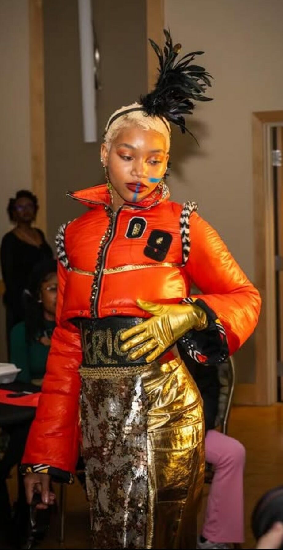 Woman with platinum blonde hair and black feathered headpiece, wearing a bright orange jacket, gold gloves, and patterned pants, in an indoor setting.