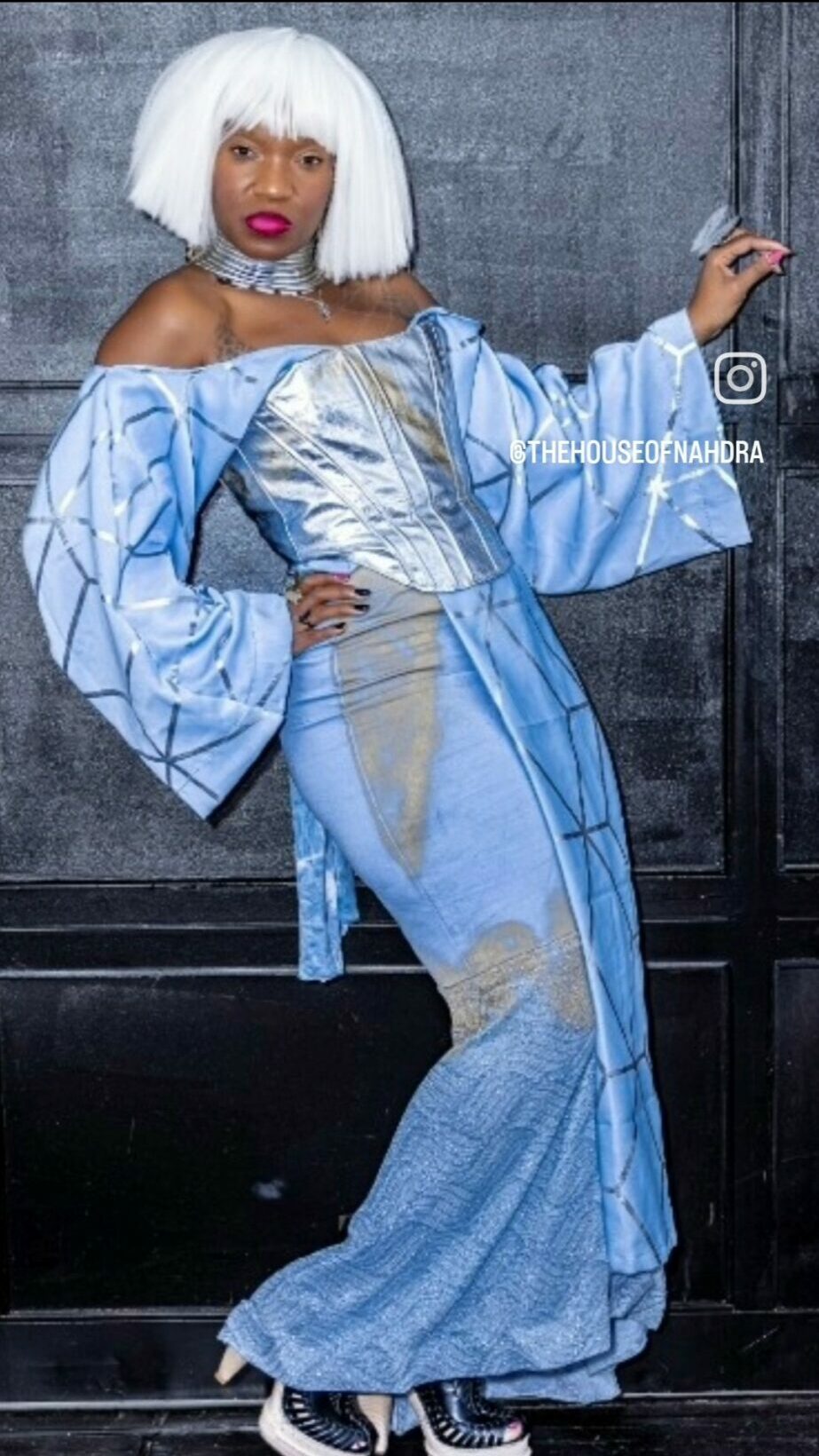 Woman with platinum blonde wig in metallic blue dress and oversized sleeves, standing against a dark background.