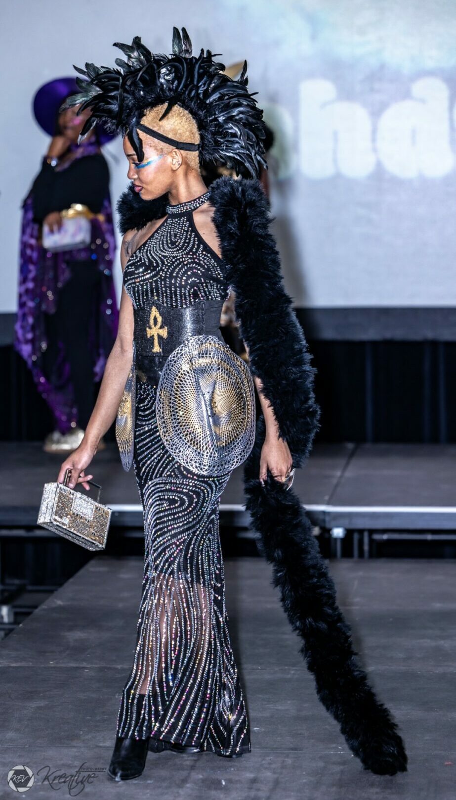 Person in elaborate costume with black feathered headdress and long tail, holding a clutch, on stage.