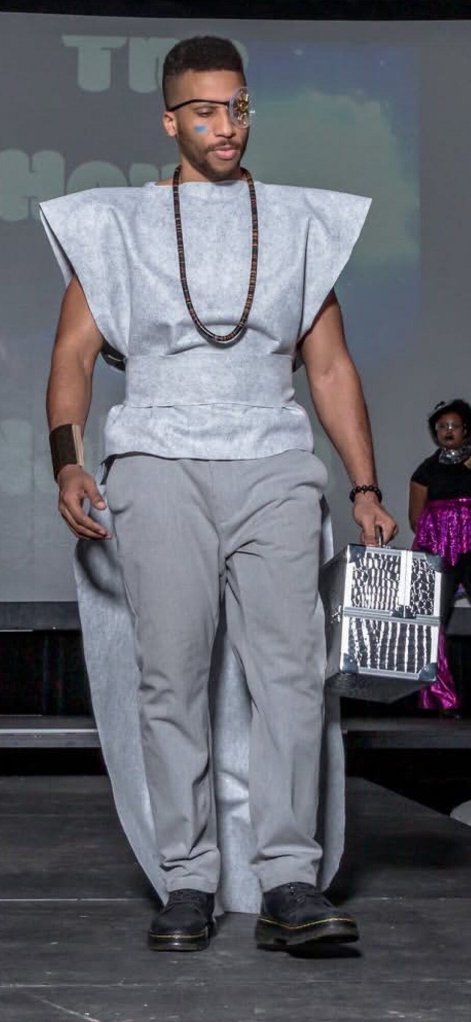 Man walking on stage wearing traditional attire, glasses, and a beaded necklace, holding a basket, with a woman in the background.