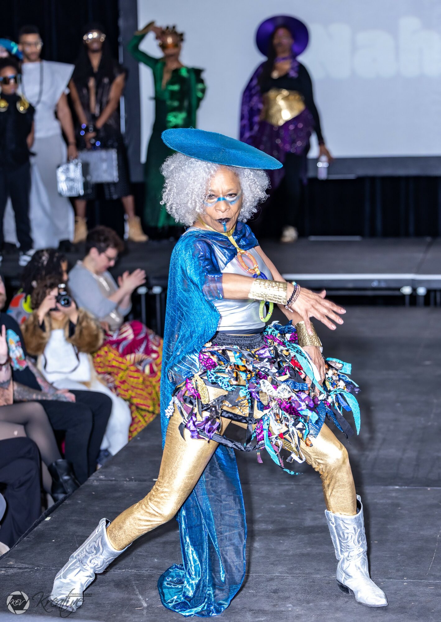 Person dressed in colorful, eclectic costume with a large blue hat, curly gray wig, and shiny boots, walking on stage.