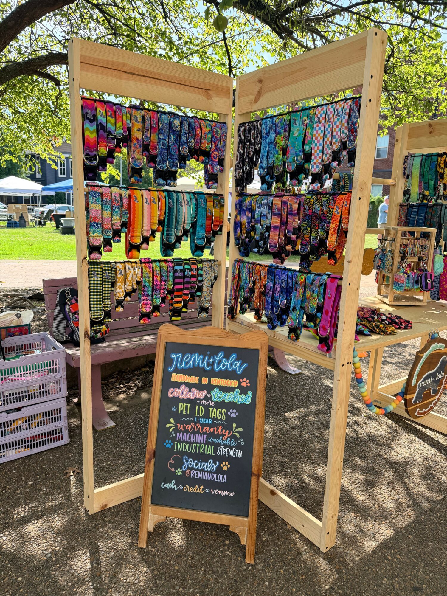 Outdoor yarn and craft booth with colorful yarns on display, a chalkboard sign, and trees in the background.