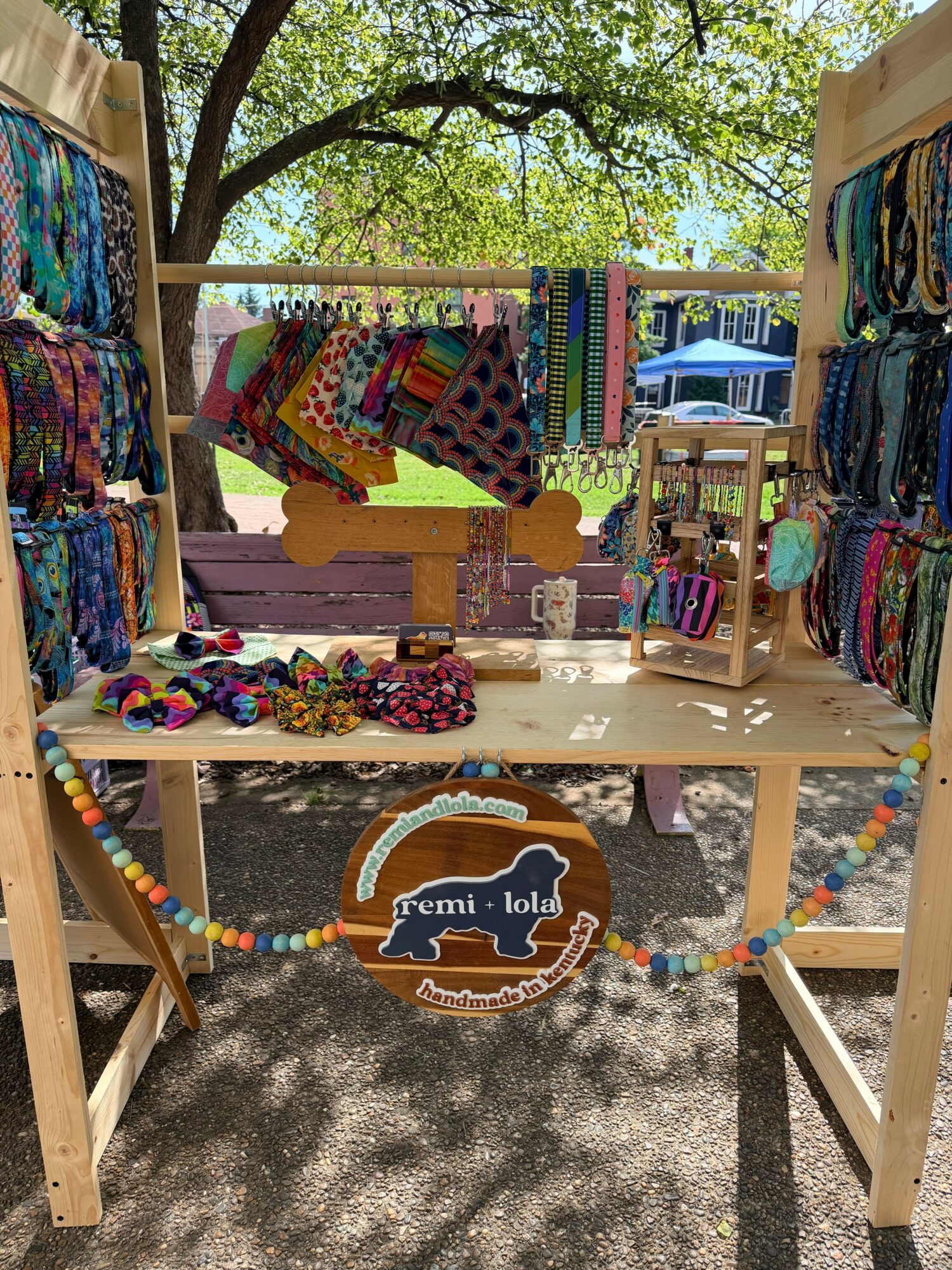 Outdoor market stall with colorful fabric, accessories, and a sign, shaded by trees, on a sunny day.