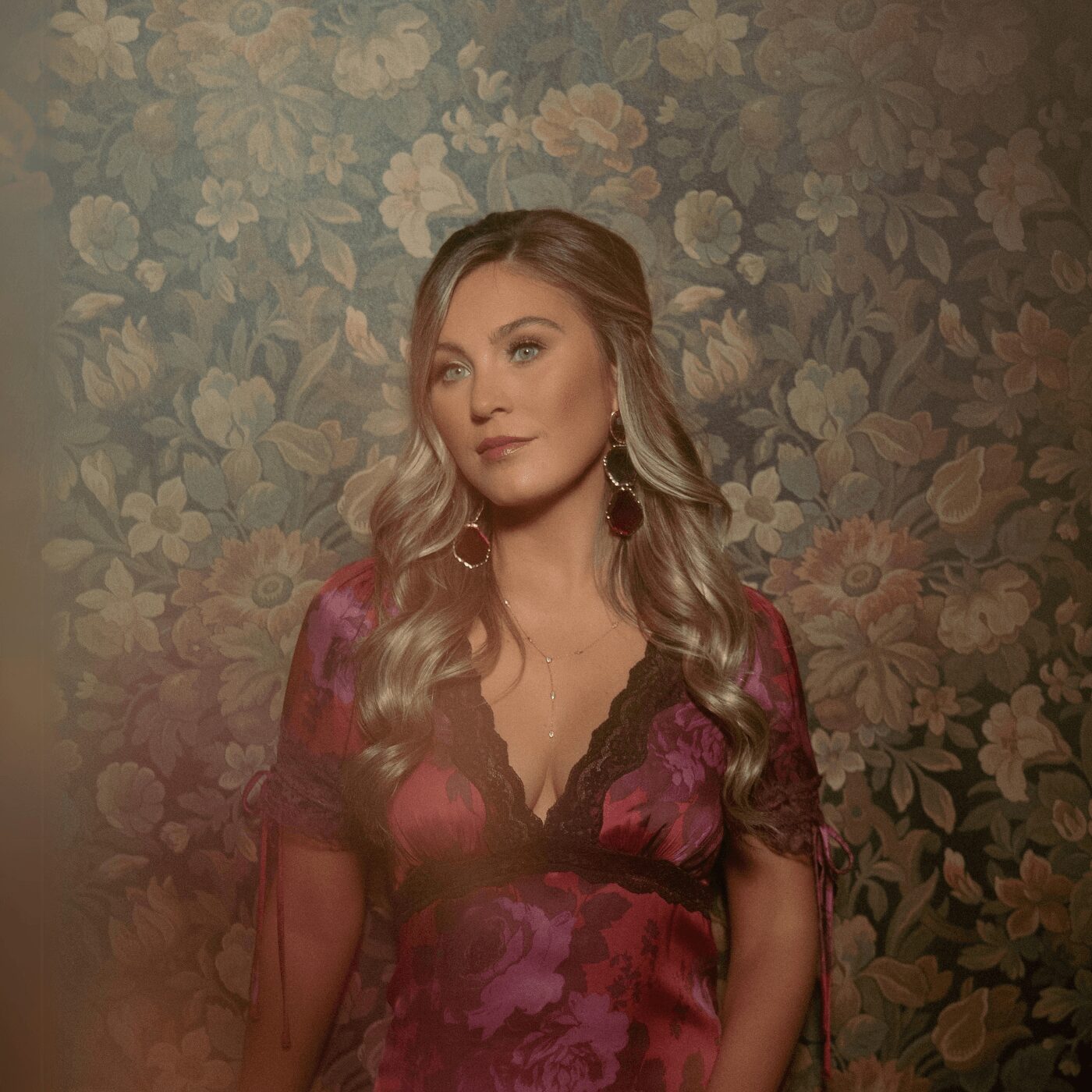 Woman with long wavy blonde hair wearing a floral dress, standing against a floral wallpaper background.