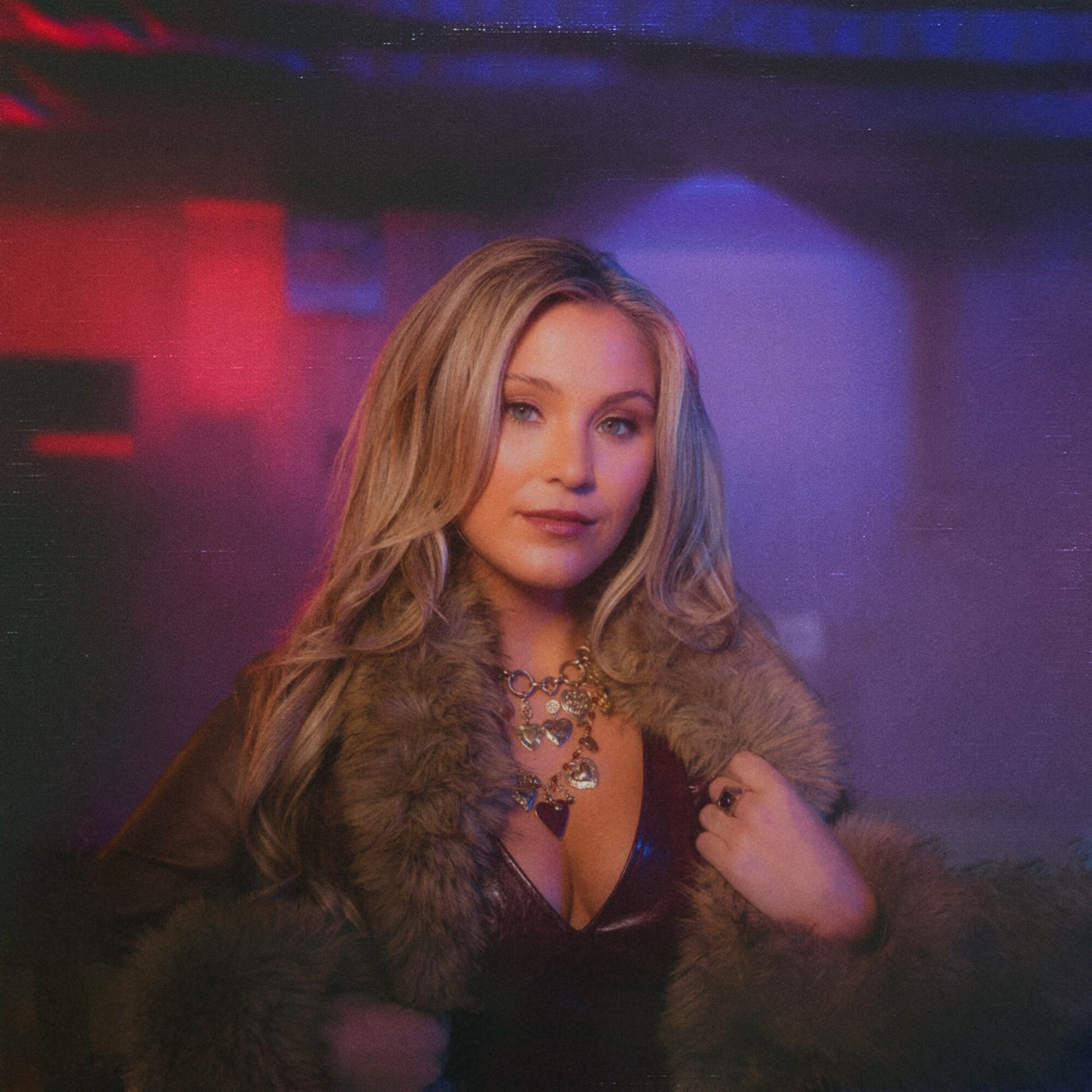 Woman with long blonde hair wearing a fur coat and jewelry, standing in a dimly lit environment with colorful lights.