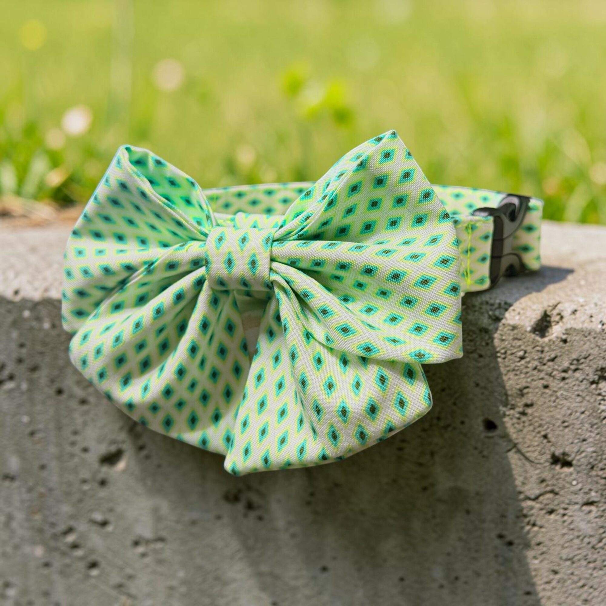 A green and blue patterned fabric bow on a concrete surface with grass in the background.