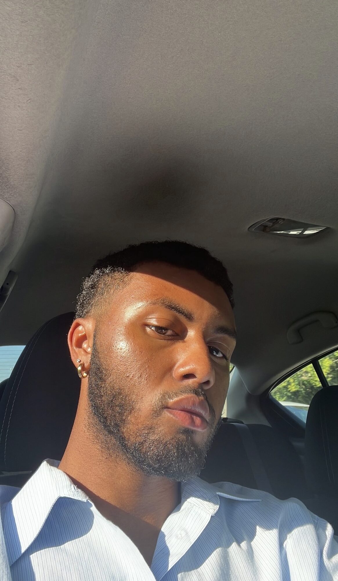 Young man with short hair and beard sitting in car, wearing a white shirt, sunlight on face, looking at camera.