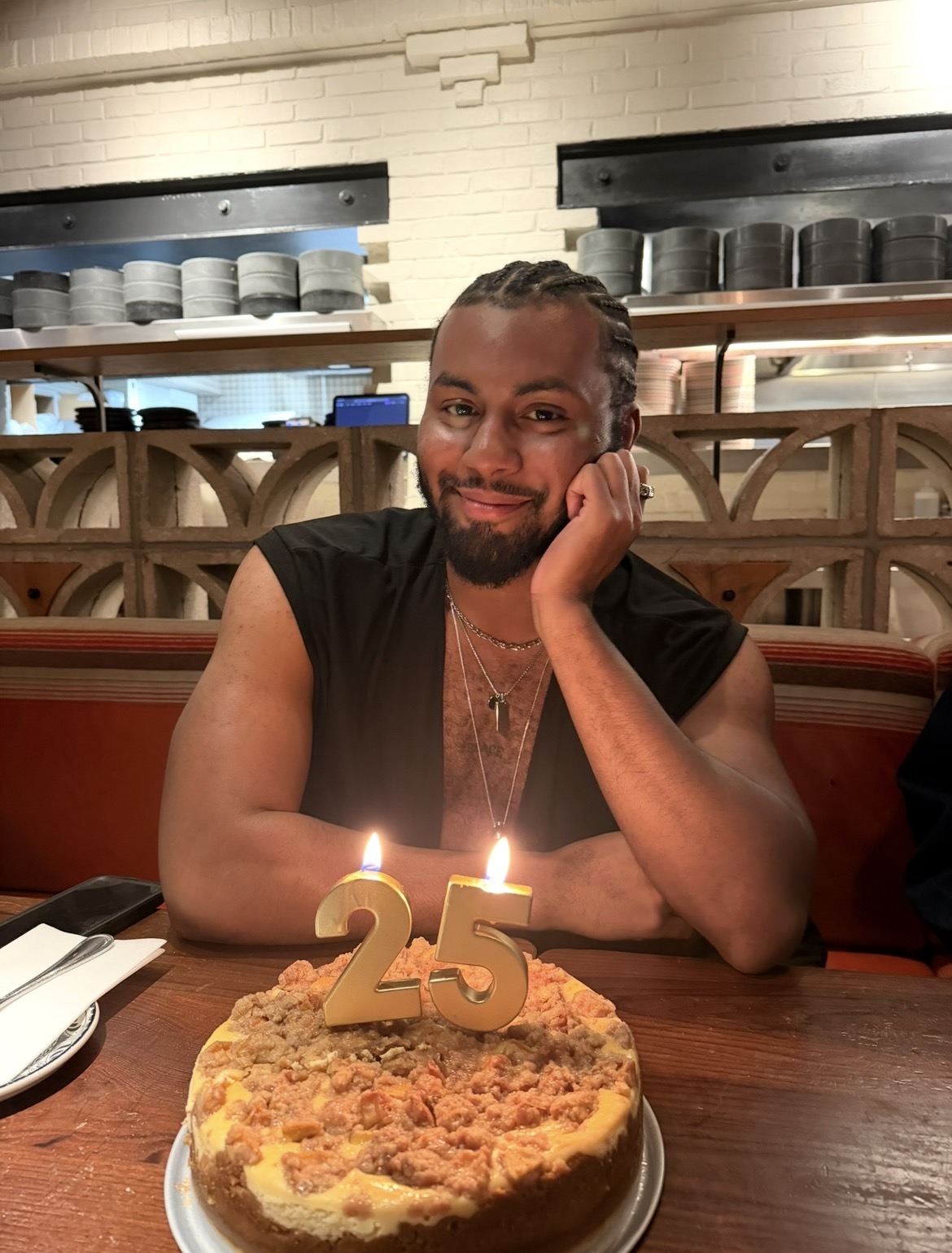 Person with braided hair and beard celebrating birthday with a cake and candles, smiling in a restaurant setting.