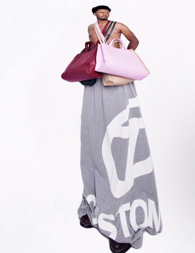 Person holding multiple large bags, wearing a long skirt with a large graphic design, standing against a white background.