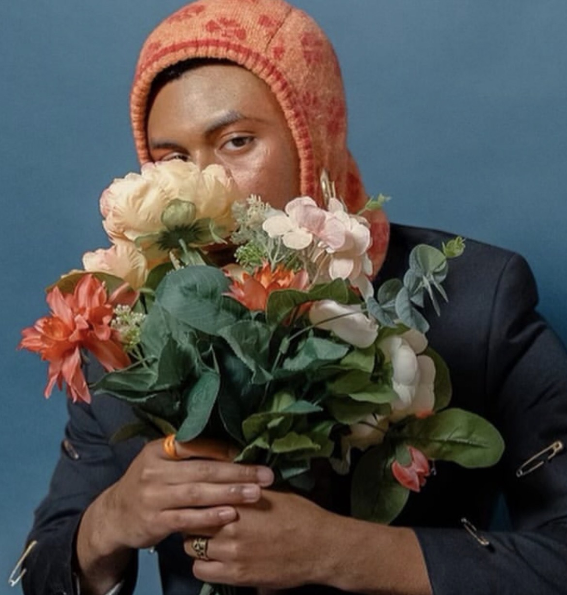 Person holding a bouquet of colorful flowers, wearing a patterned orange hat and a dark jacket, against a blue background.