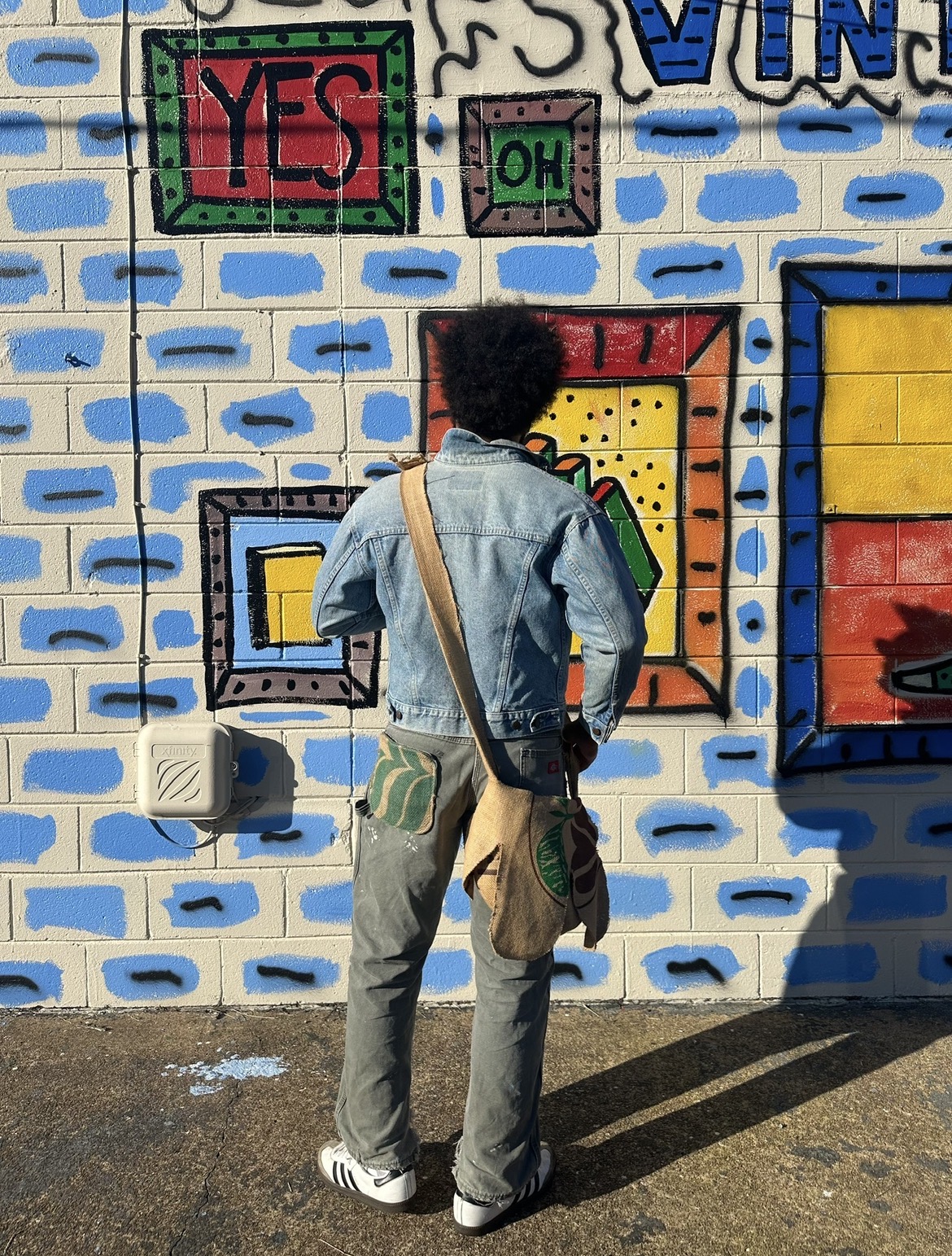 Person with curly hair, wearing a denim jacket and gray pants, stands in front of a colorful graffiti wall.