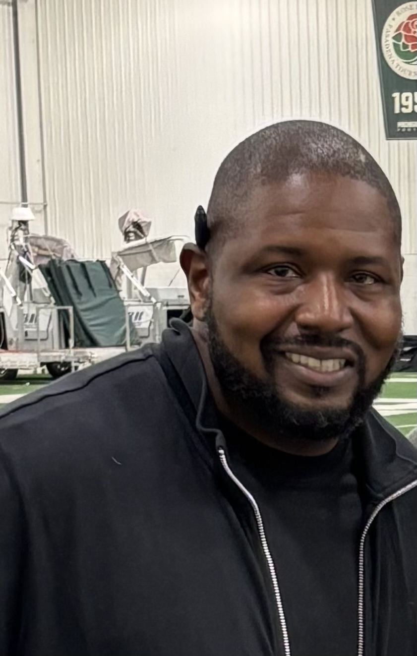 Man with short hair and beard smiling, wearing a black jacket, indoors with a sports stadium background.