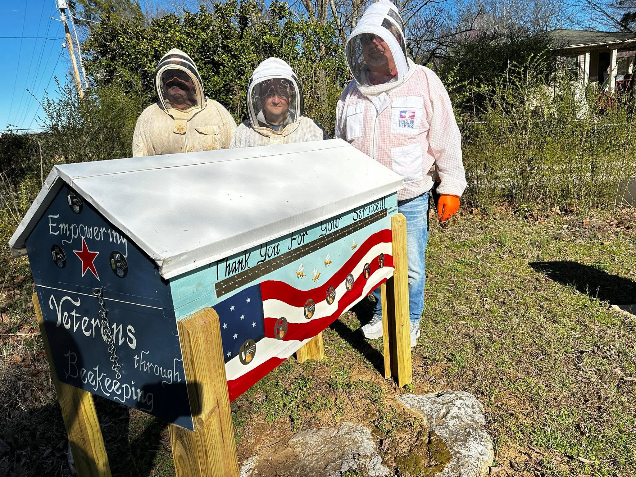 Three people in protective suits and veils stand outdoors near a decorated box with an American flag design, grass, and trees.