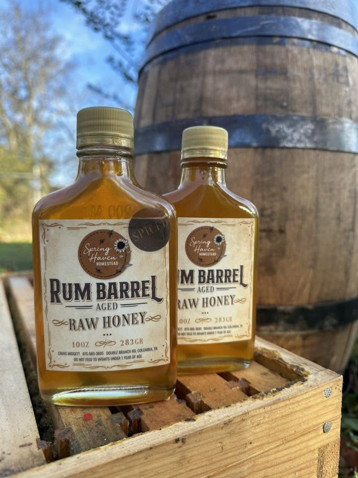 Two bottles of Rum Barrel raw honey placed on a wooden crate outdoors with a large barrel in the background.