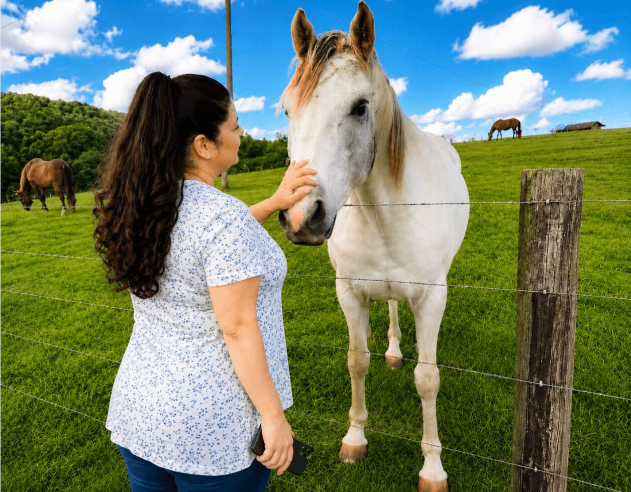 Woman petting a white horse through a fence in a green field with blue sky and clouds.