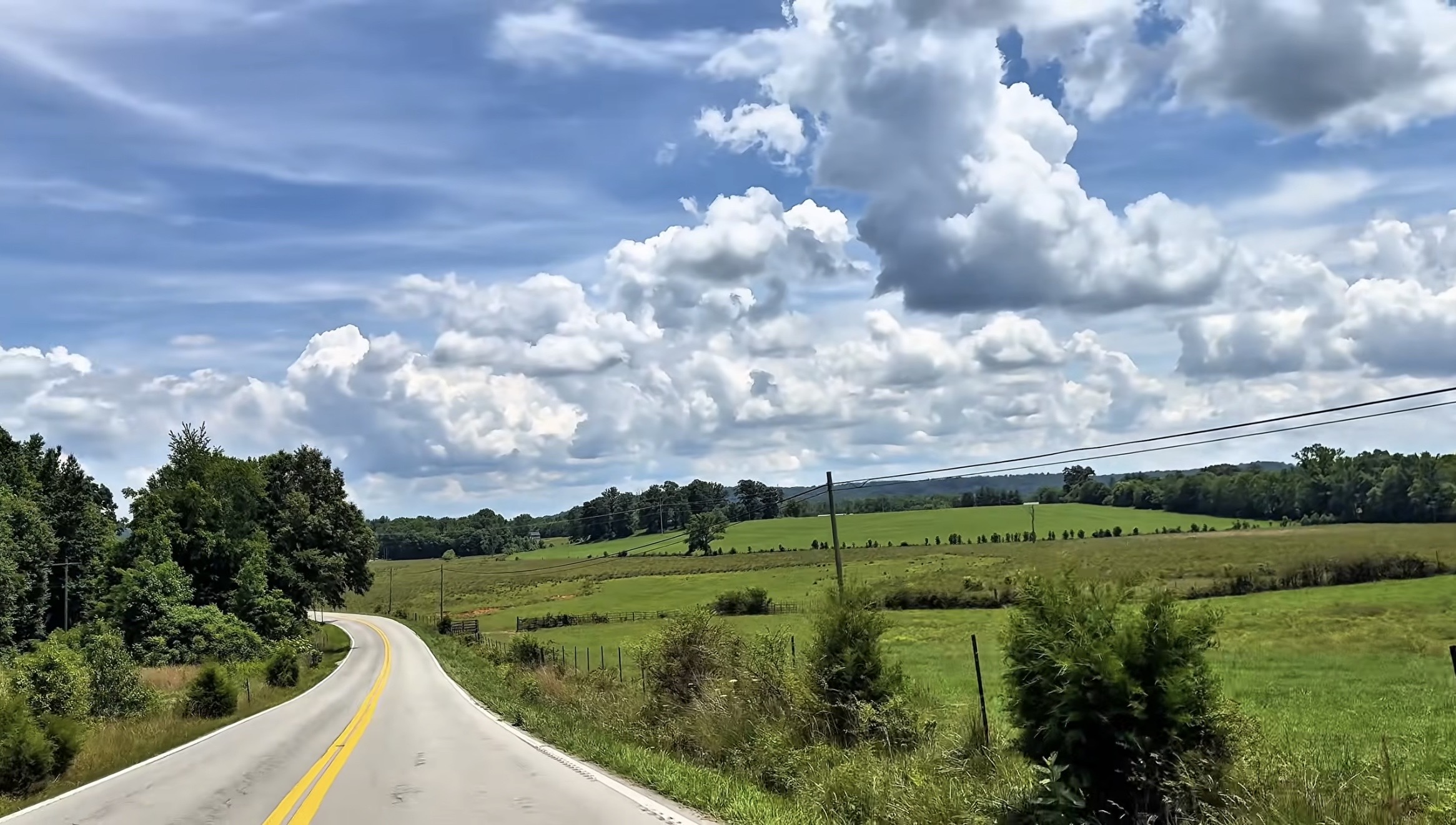 Scenic rural landscape with a winding road, green fields, trees, and a partly cloudy sky.