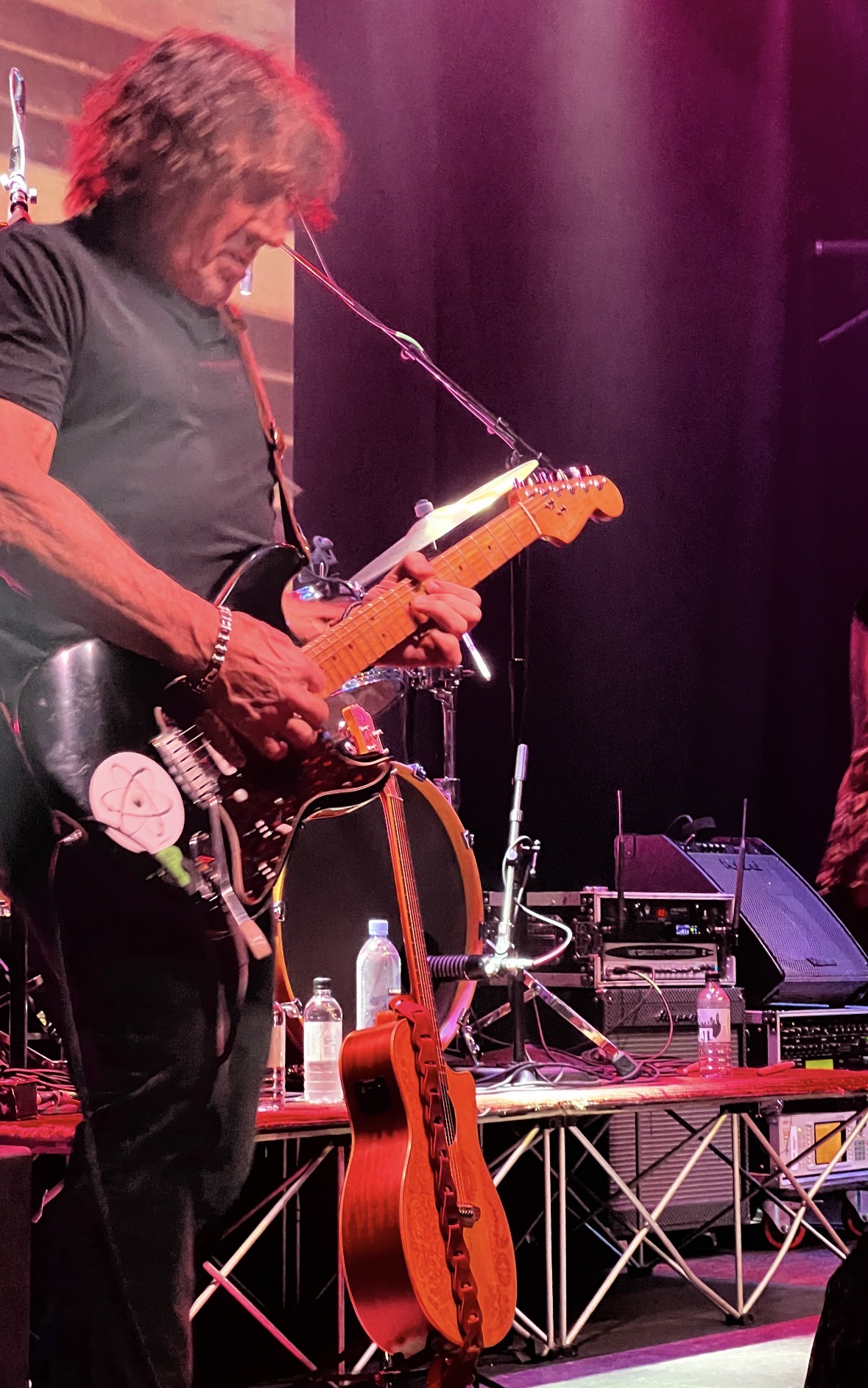 Musician playing electric guitar on stage with pink and purple lighting, equipment, and a red acoustic guitar nearby.