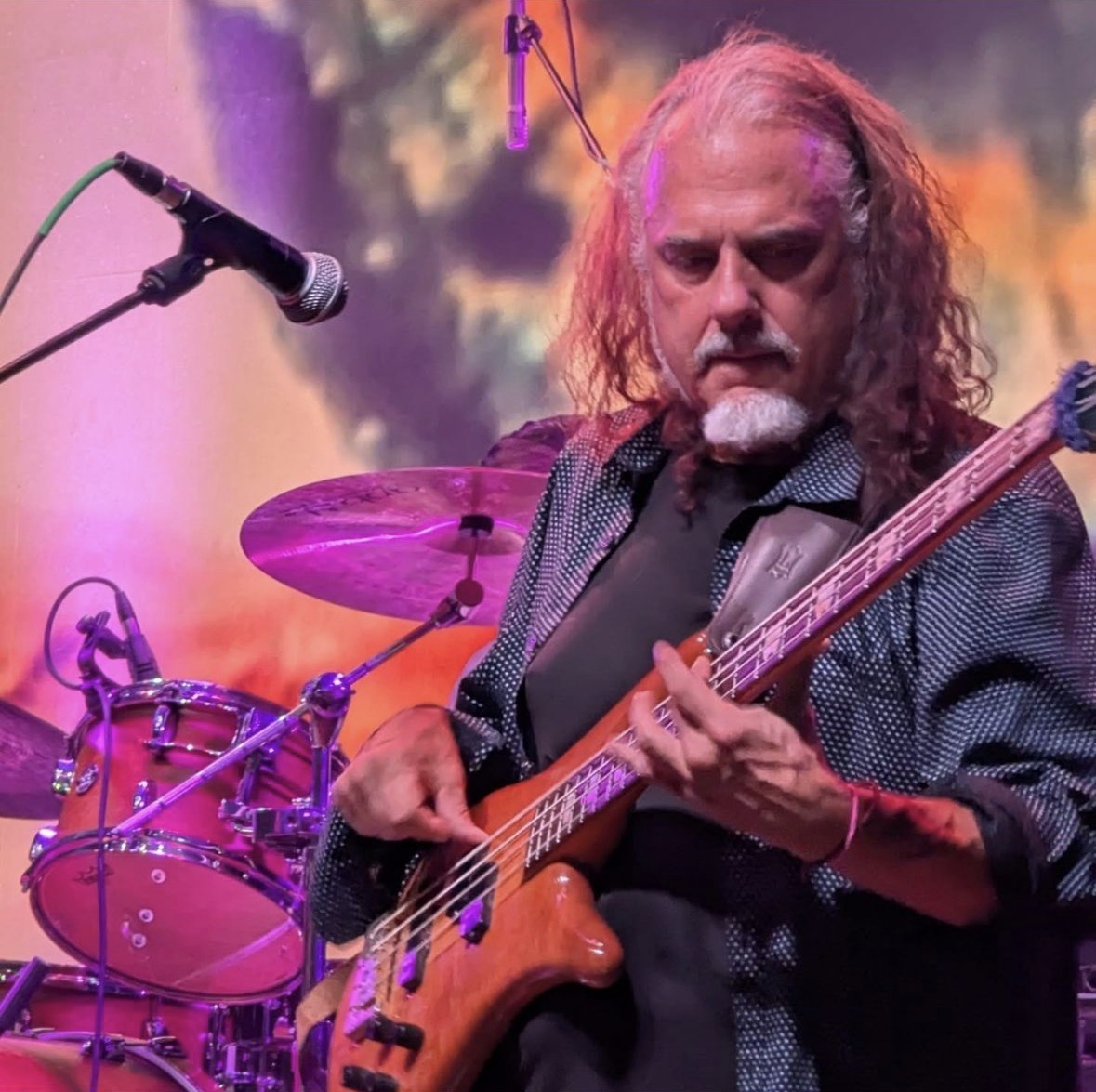 Musician playing bass guitar on stage with drums and colorful background, focused on performance.
