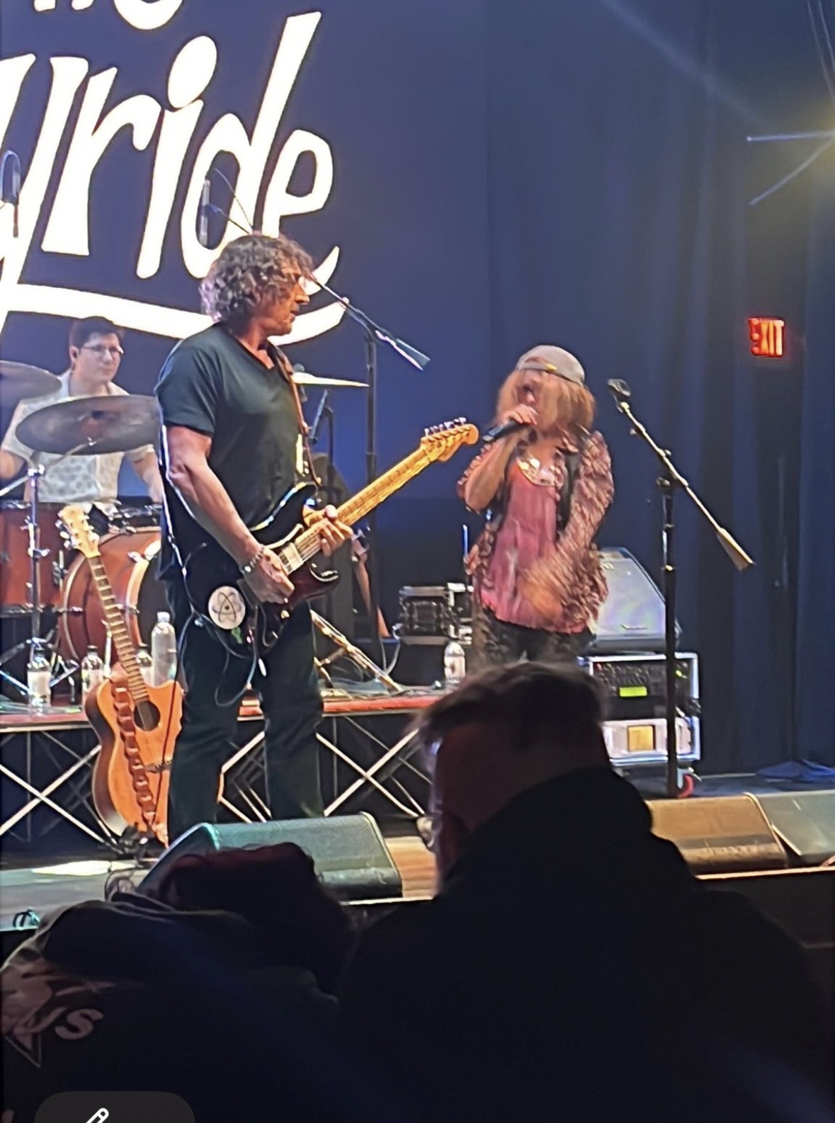 Musicians perform on stage with a guitarist and singer, stage lights, and an audience in foreground, with a partially visible word 'ride' in background.