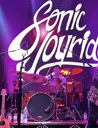 A drummer performing on stage with a large purple backdrop displaying the text 'Sonic Flourish'.