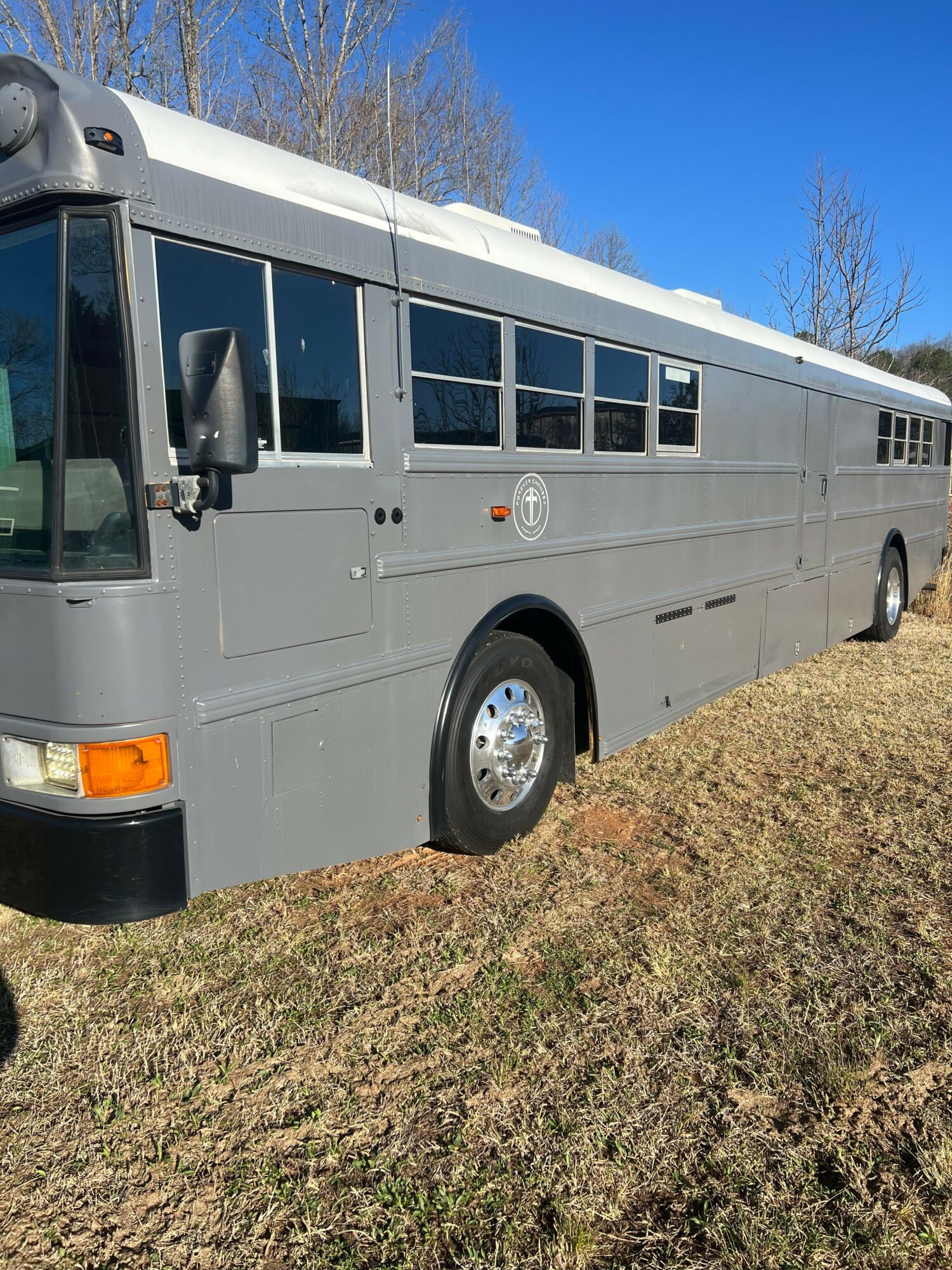 Grey bus parked on grass with clear blue sky in background, side view showing windows and wheel.