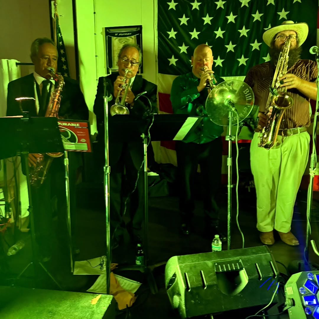 Four musicians playing saxophones on stage with a green background and American flag, with music stands and equipment visible.