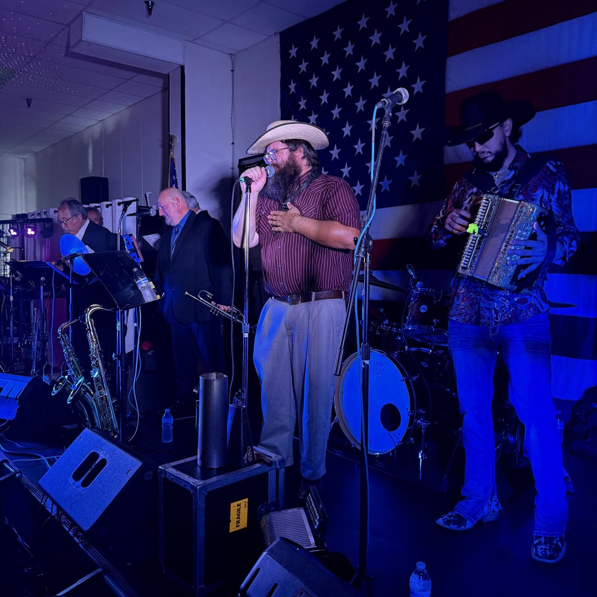 Band performs on stage with American flag backdrop, featuring a singer with a hat and musicians playing instruments.