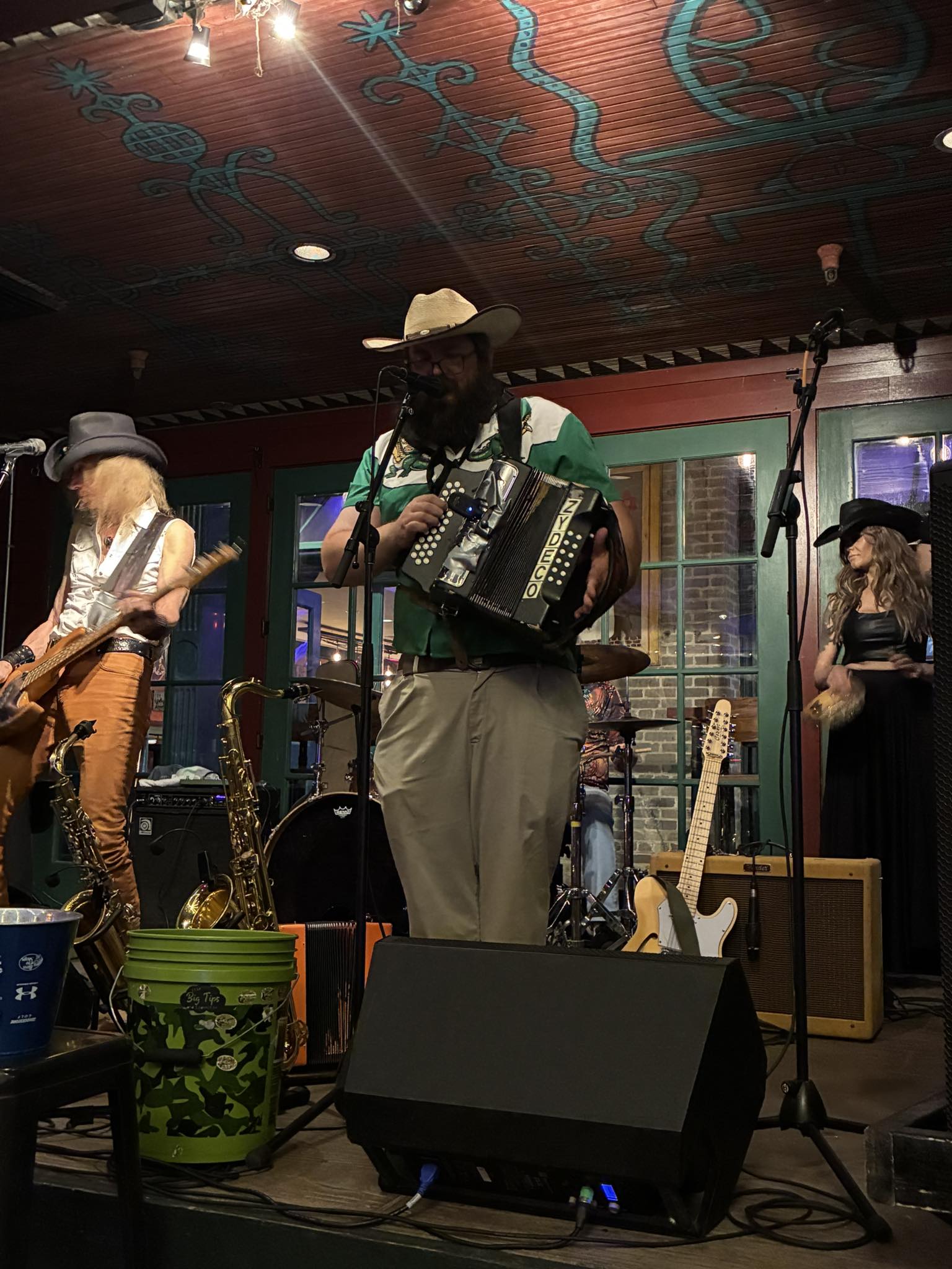 Musicians perform on stage with instruments, including an accordion, in a cozy indoor venue with colorful decor.