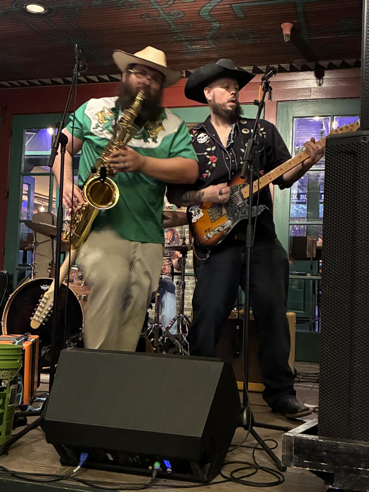 Two musicians perform on stage, one playing saxophone and the other playing guitar, with microphones and stage equipment visible.