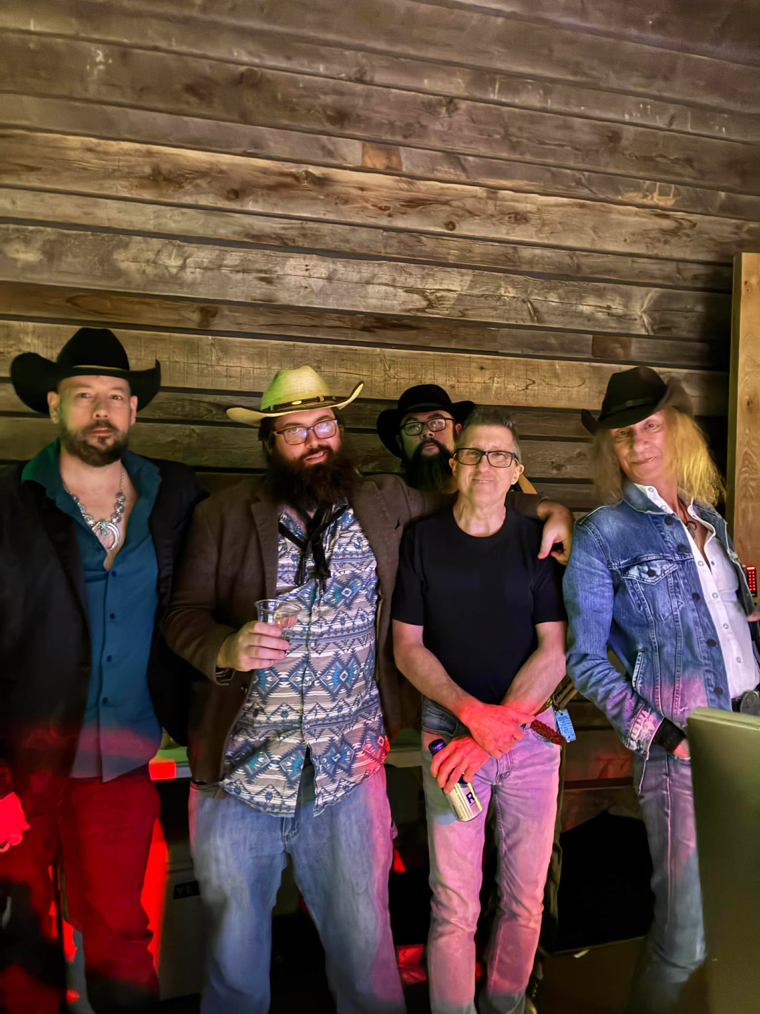 Five people standing together indoors against a wooden wall, some wearing hats and casual clothing.