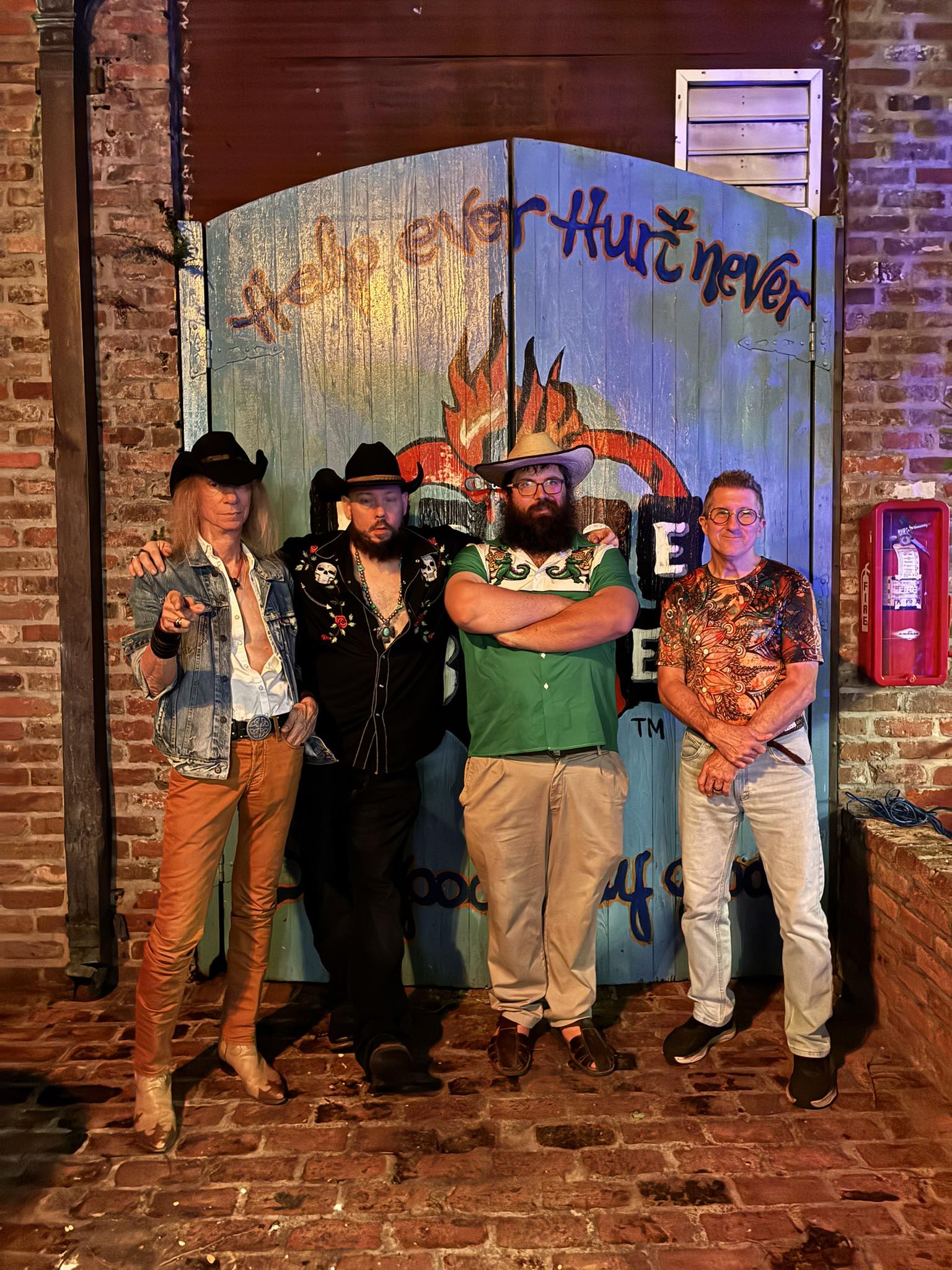 Four people stand in front of a painted backdrop with a fire and text, inside a brick-walled venue, with a fire extinguisher nearby.