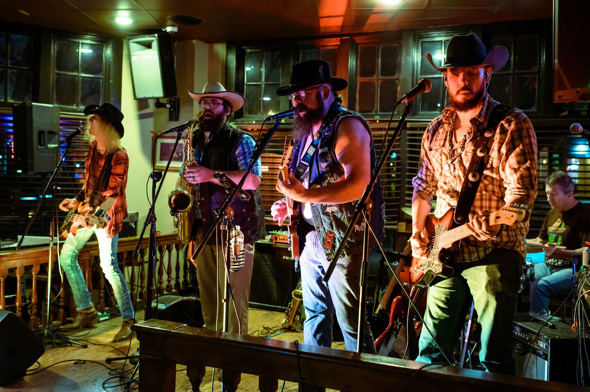 Four musicians perform on stage with instruments, wearing hats and casual clothing, in a dimly lit venue.