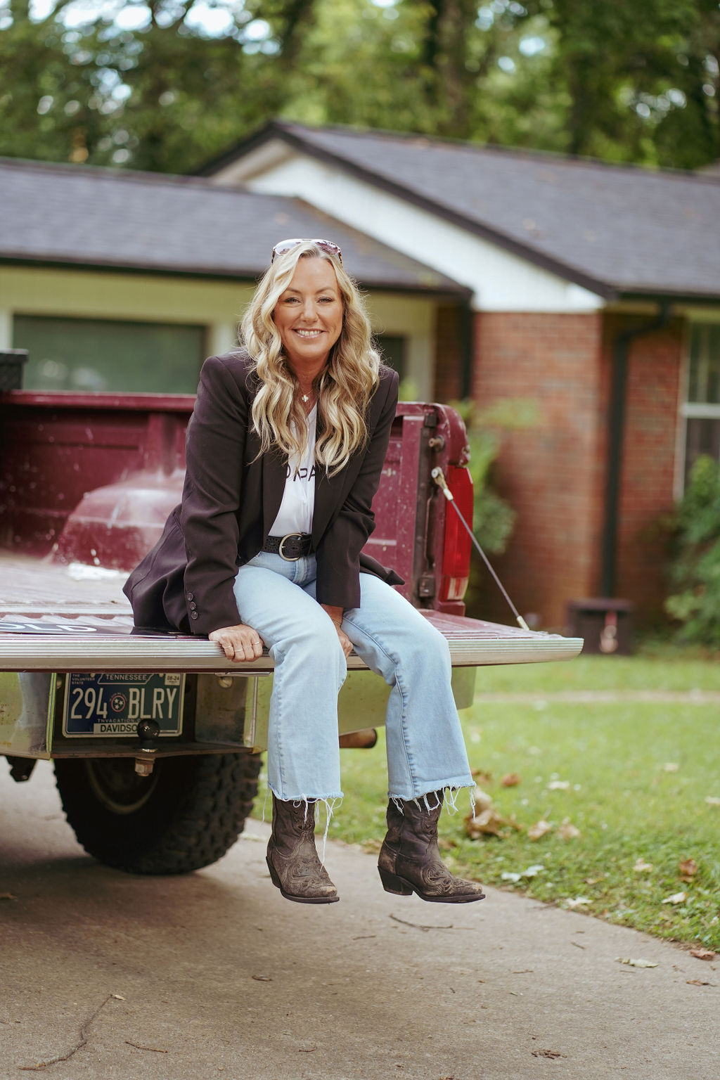 Woman with long hair sitting on a truck bed, smiling, in front of a house with trees in the background.