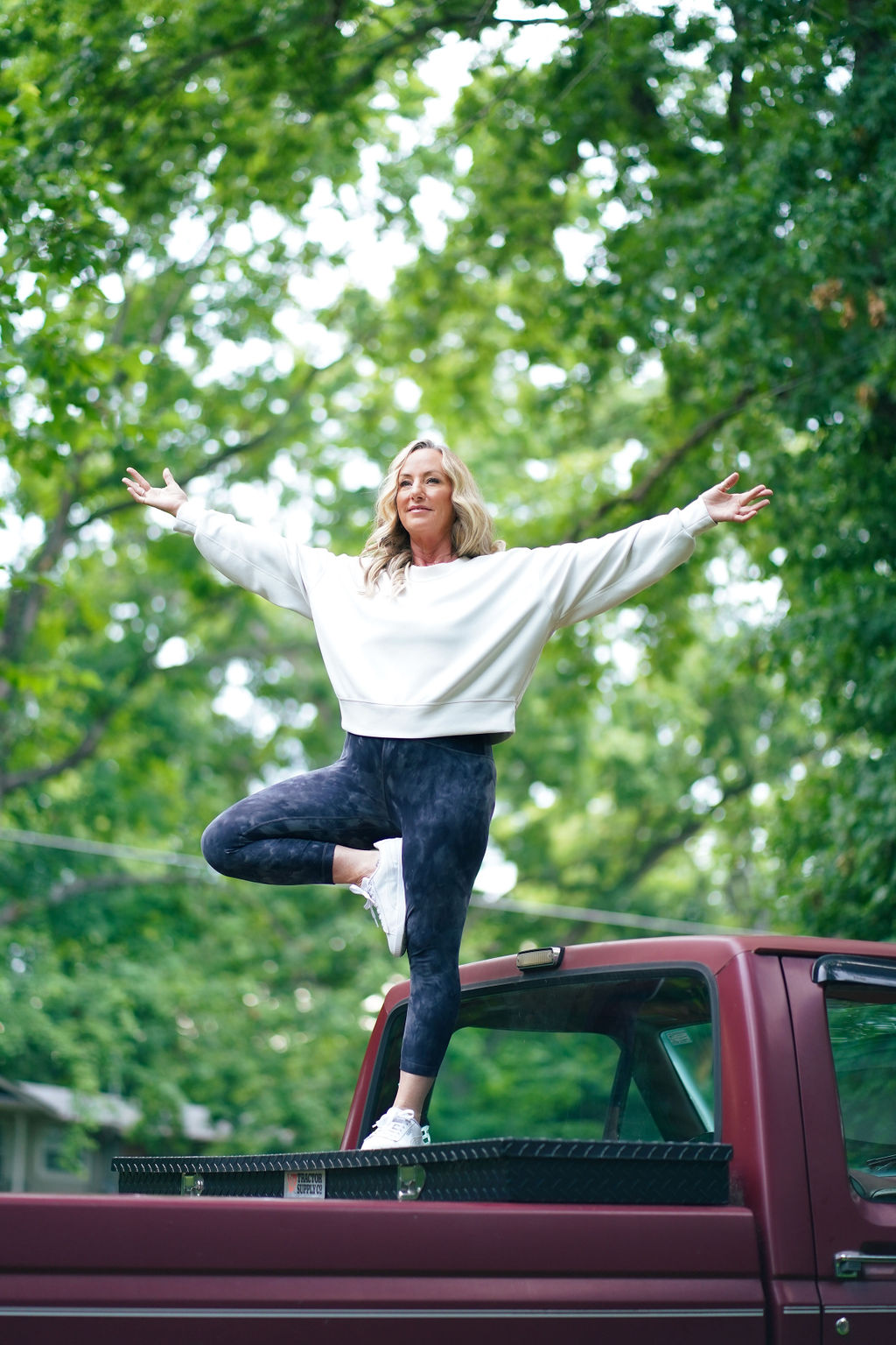 Woman standing on a vehicle's roof with arms outstretched, surrounded by green trees, smiling, wearing a white sweater and dark pants.