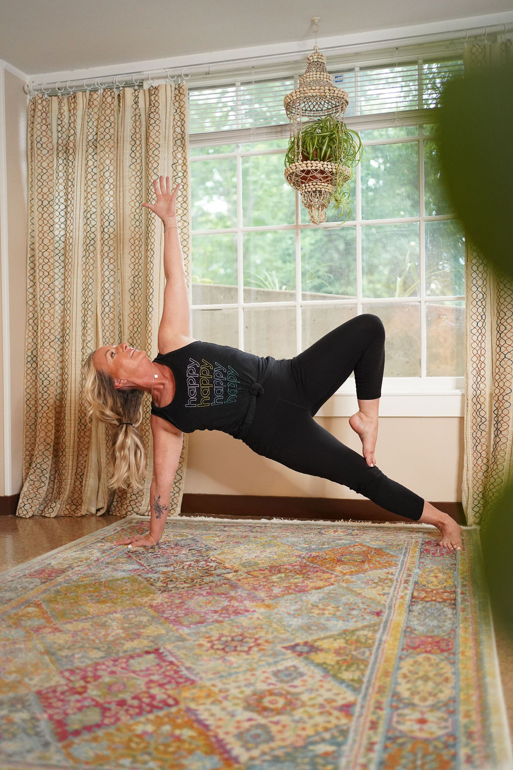 Woman practicing yoga in a living room with large window, patterned curtains, and a hanging plant chandelier.